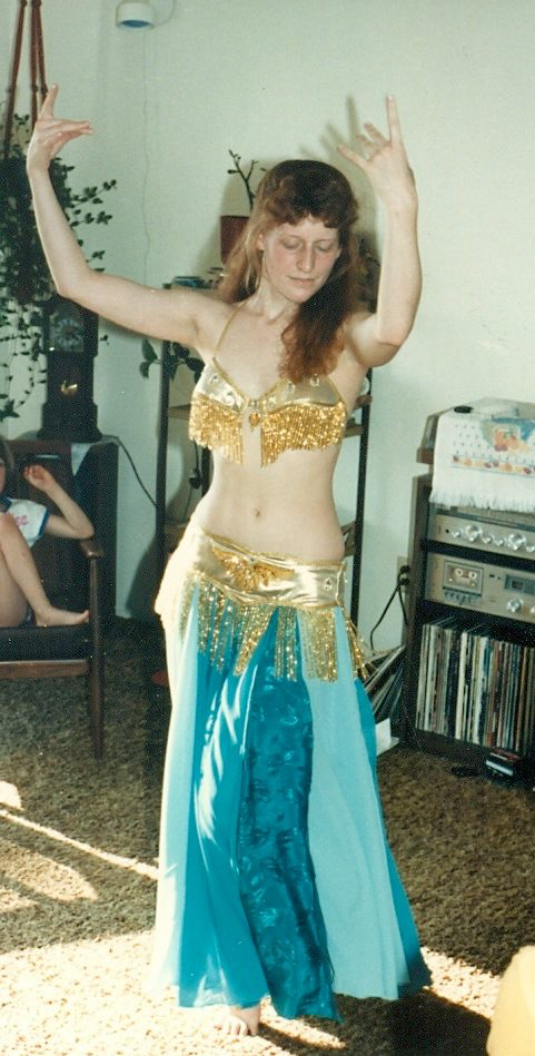 The author, age 23, dancing for friends and family at her college graduation party, wearing a gold lame bra and hip-belt decorated with bead fringe, over two layers of teal polyester dance skirts.