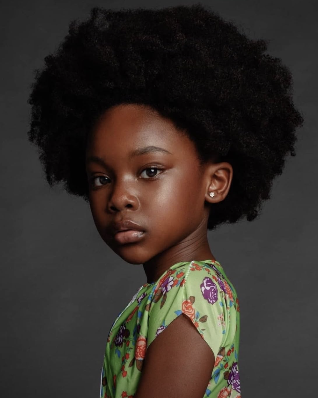 A young girl with dark skin and an afro hairstyle, wearing a green floral dress and pearl earrings, looking at the camera with a neutral expression against a dark background.