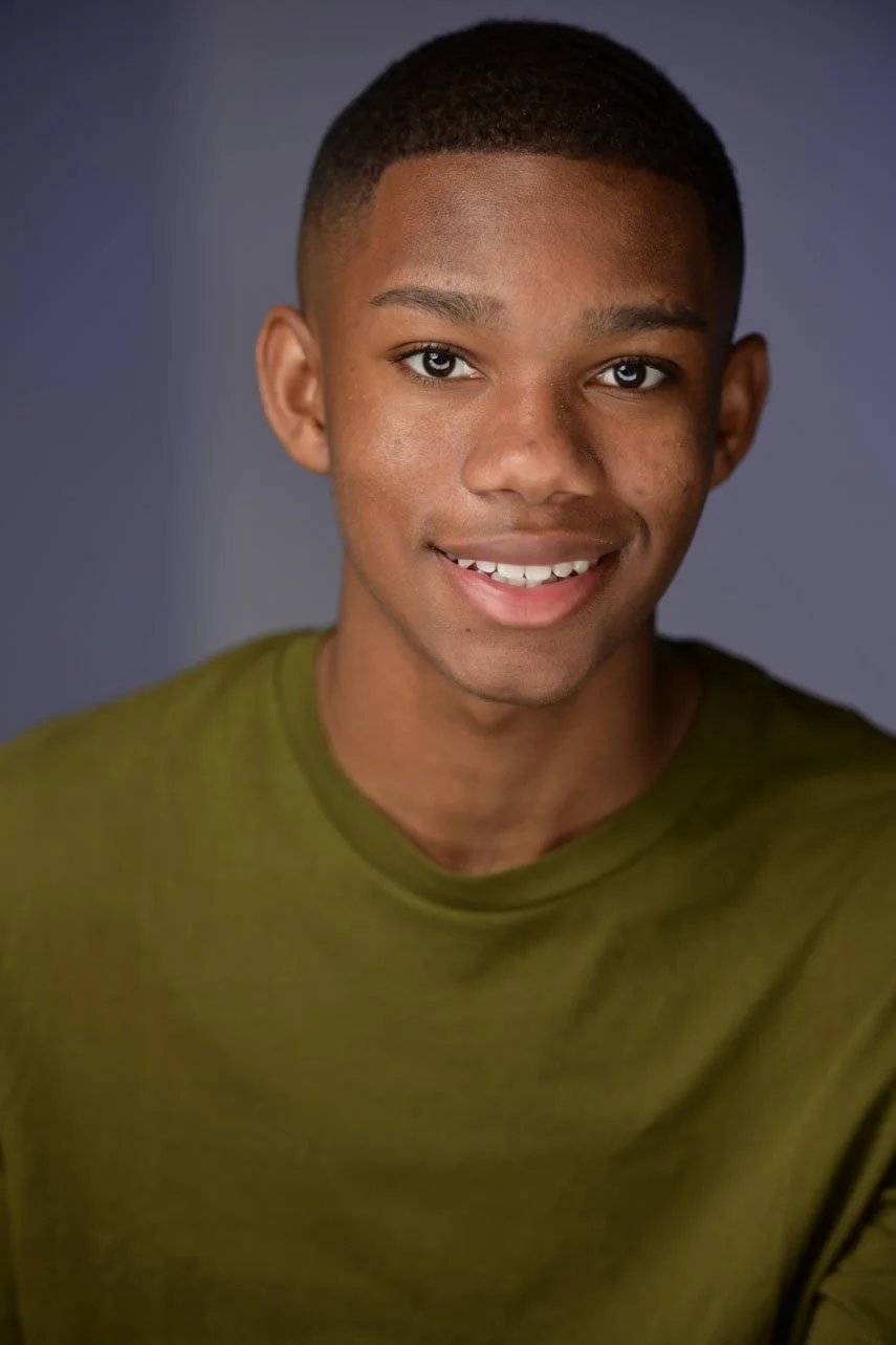 A smiling young boy with short hair, wearing an orange shirt and a distressed denim jacket, standing against a light blue background.