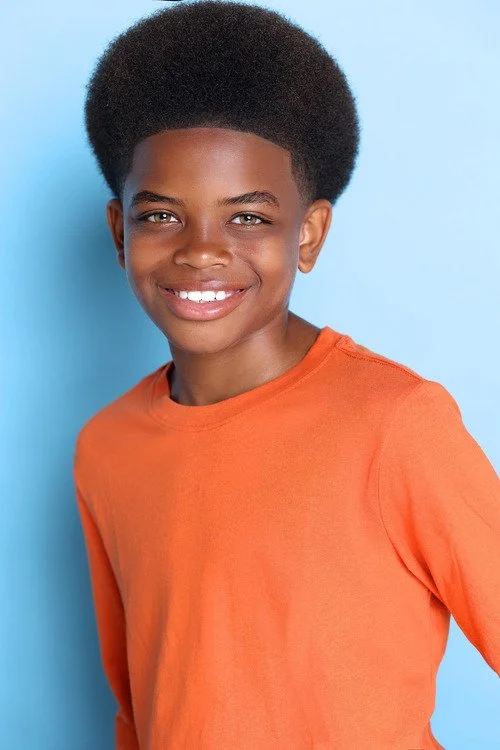 A smiling young boy with dark skin, wearing an orange long-sleeve shirt, standing against a light blue background.