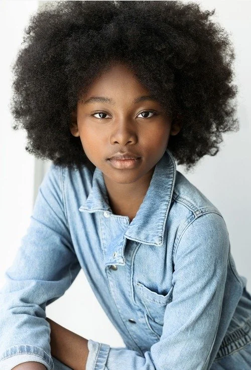 A young woman with curly black hair wearing a light blue denim jacket, looking directly into the camera.
