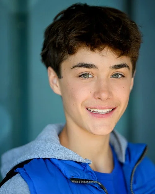 A young person with short brown hair smiling, wearing a blue jacket and a gray hoodie.