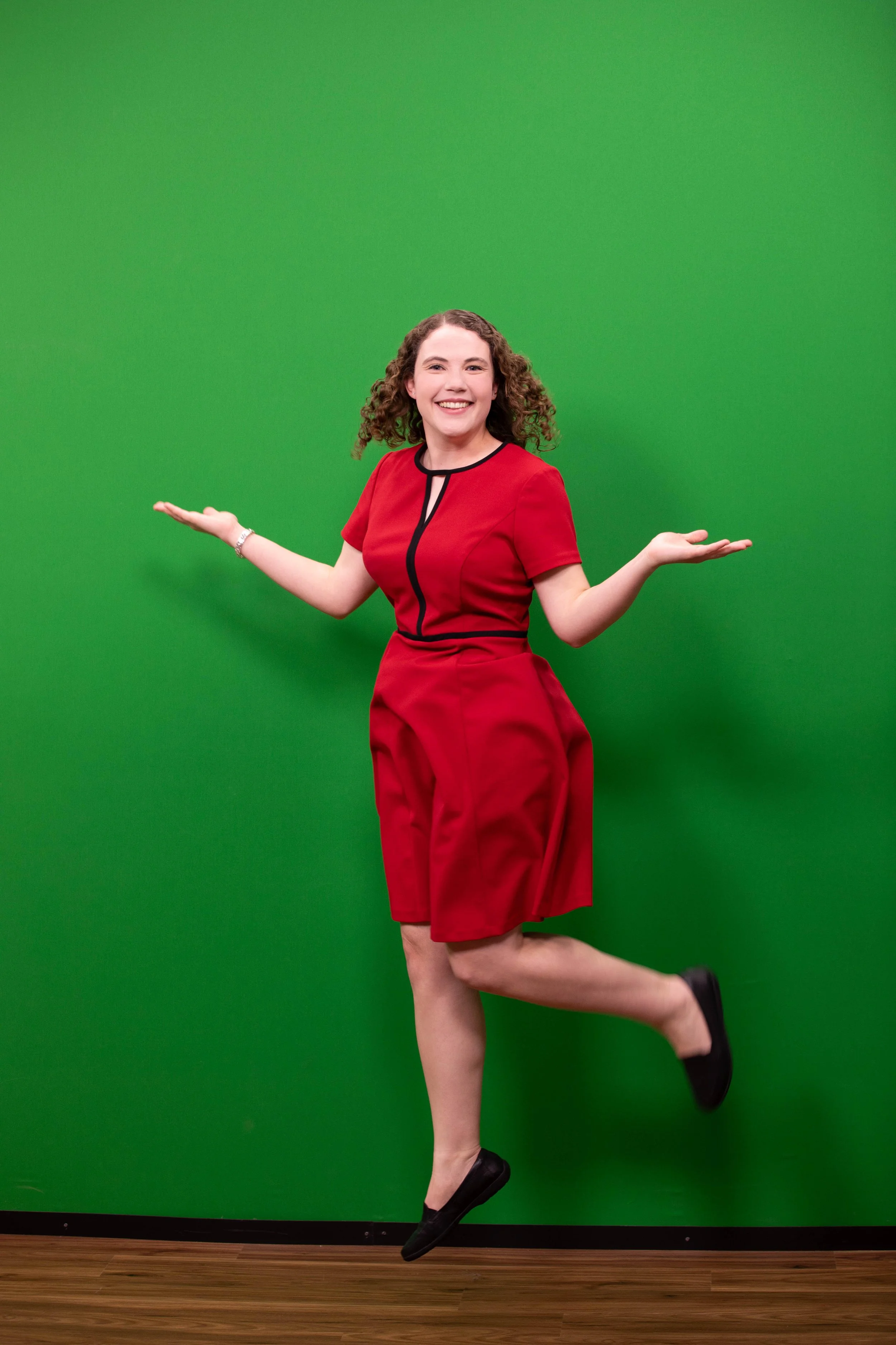 A woman with curly hair wearing a red dress with black accents, smiling and posing with arms raised, against a green background.