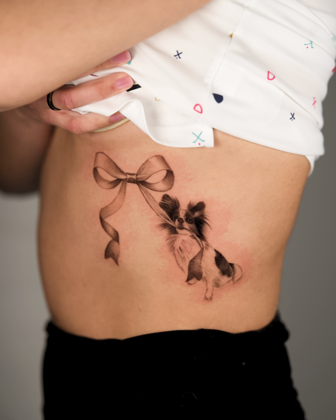 Inspired by the iconic Coppertone sunscreen ad! Fine line realism dog tattoo with ribbon bow. Placed on the ribs.