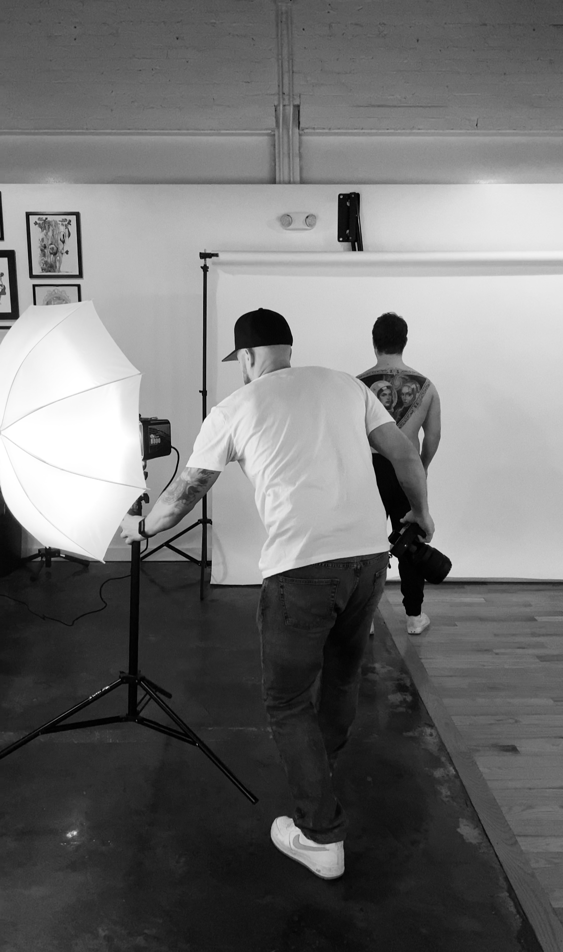 Photographer adjusting lights and camera in a studio, with a model standing in front of a white backdrop, showcasing tattoos on his back.