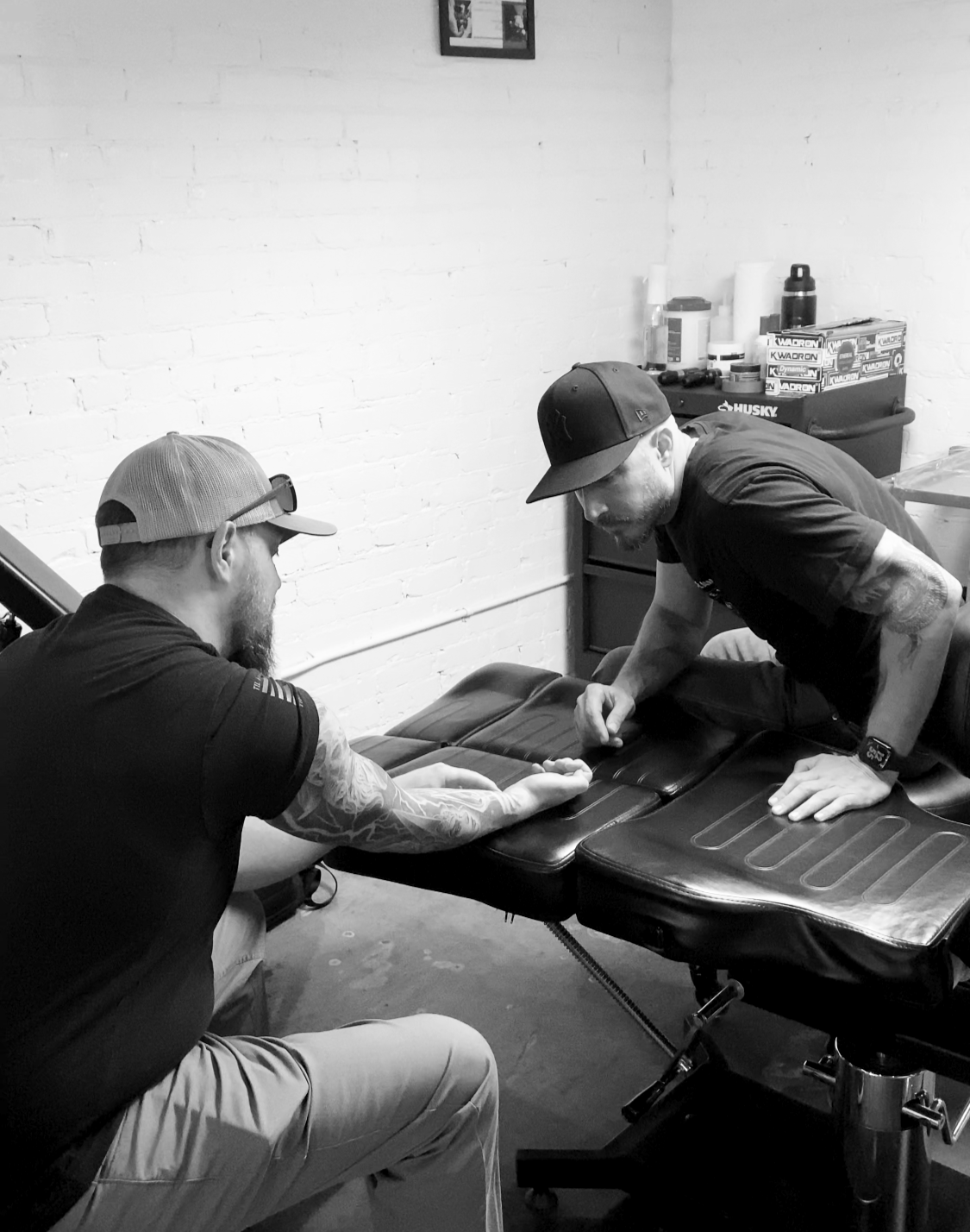 Two men in casual clothes and baseball caps, one with tattoos, are engaged in a handshake or arm-wrestling on a padded table in a room with a white brick wall. There are shelves with bottles and boxes in the background.