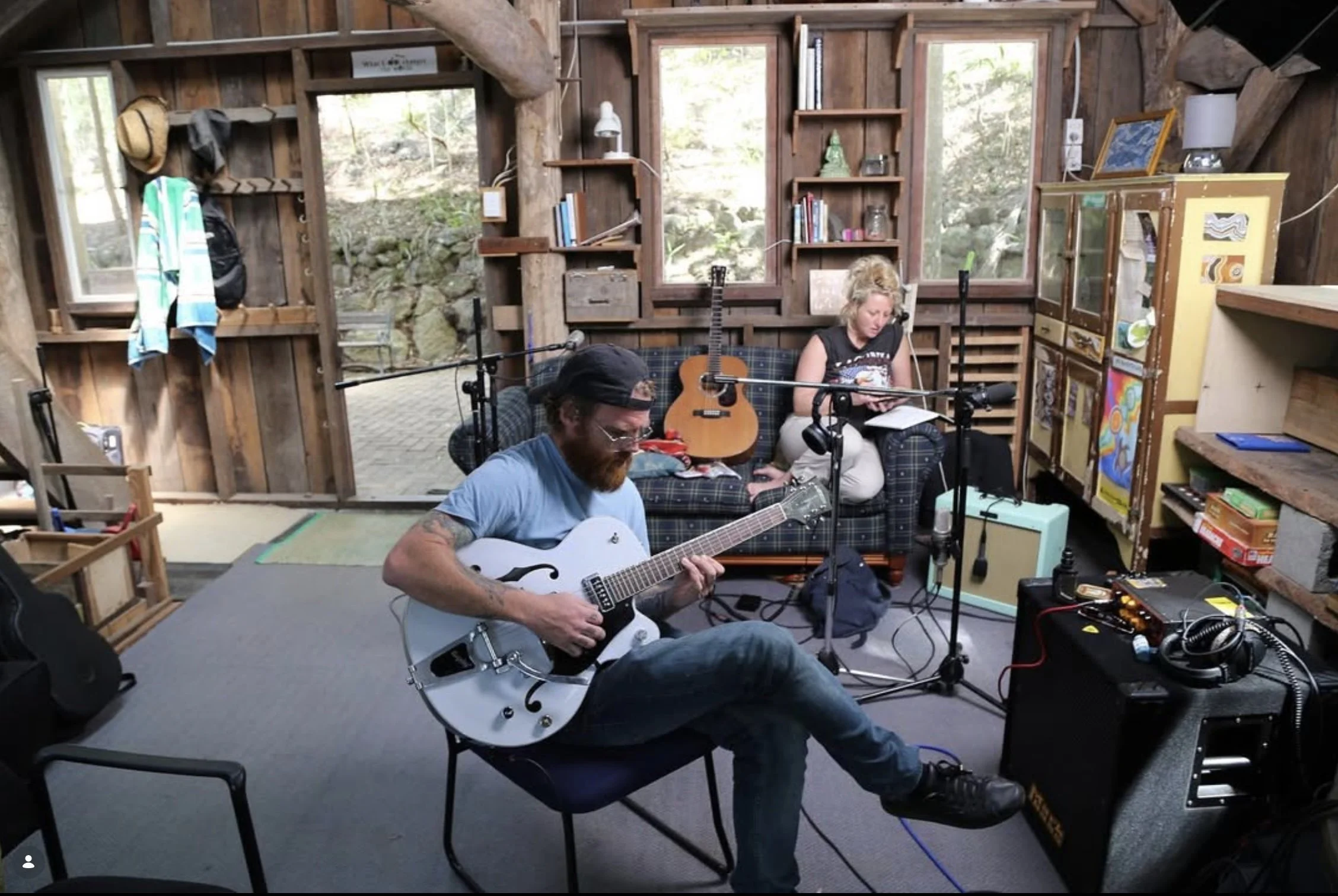 Musicians practicing in a cozy, rustic cabin with guitars, microphones, and sound equipment, surrounded by wood walls, shelves, and windows letting in natural light.