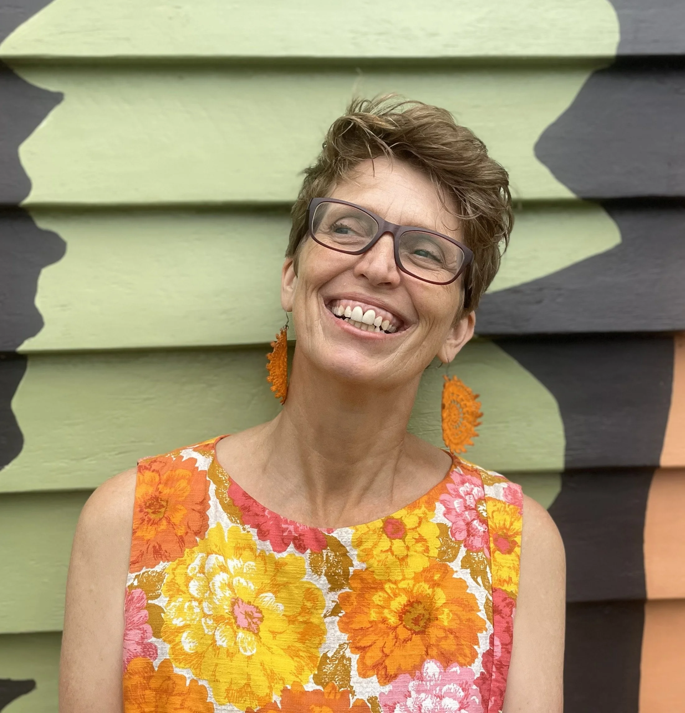 A woman with short light brown hair, wearing green glasses, pink earrings, and a pink floral dress, smiling at the camera.