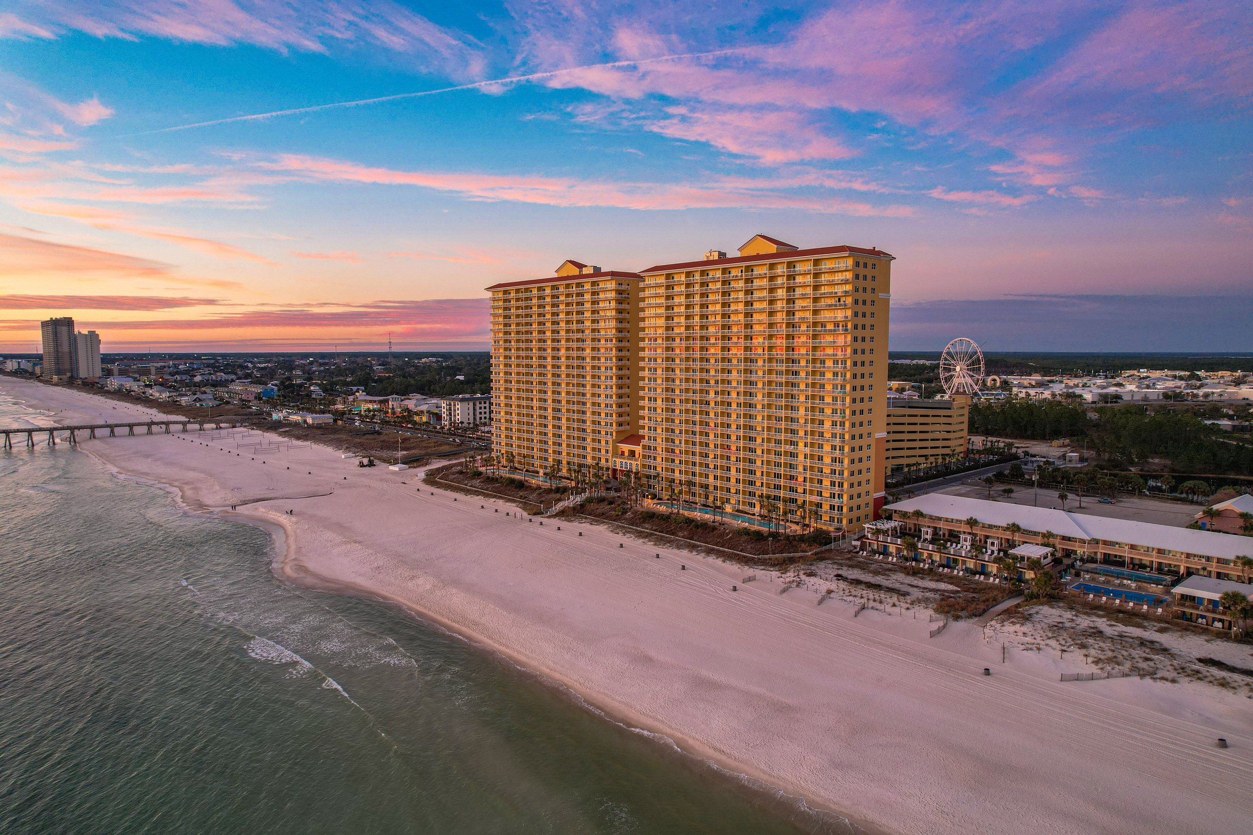 A high-rise hotel building on the beach during sunset with a Ferris wheel in the background.