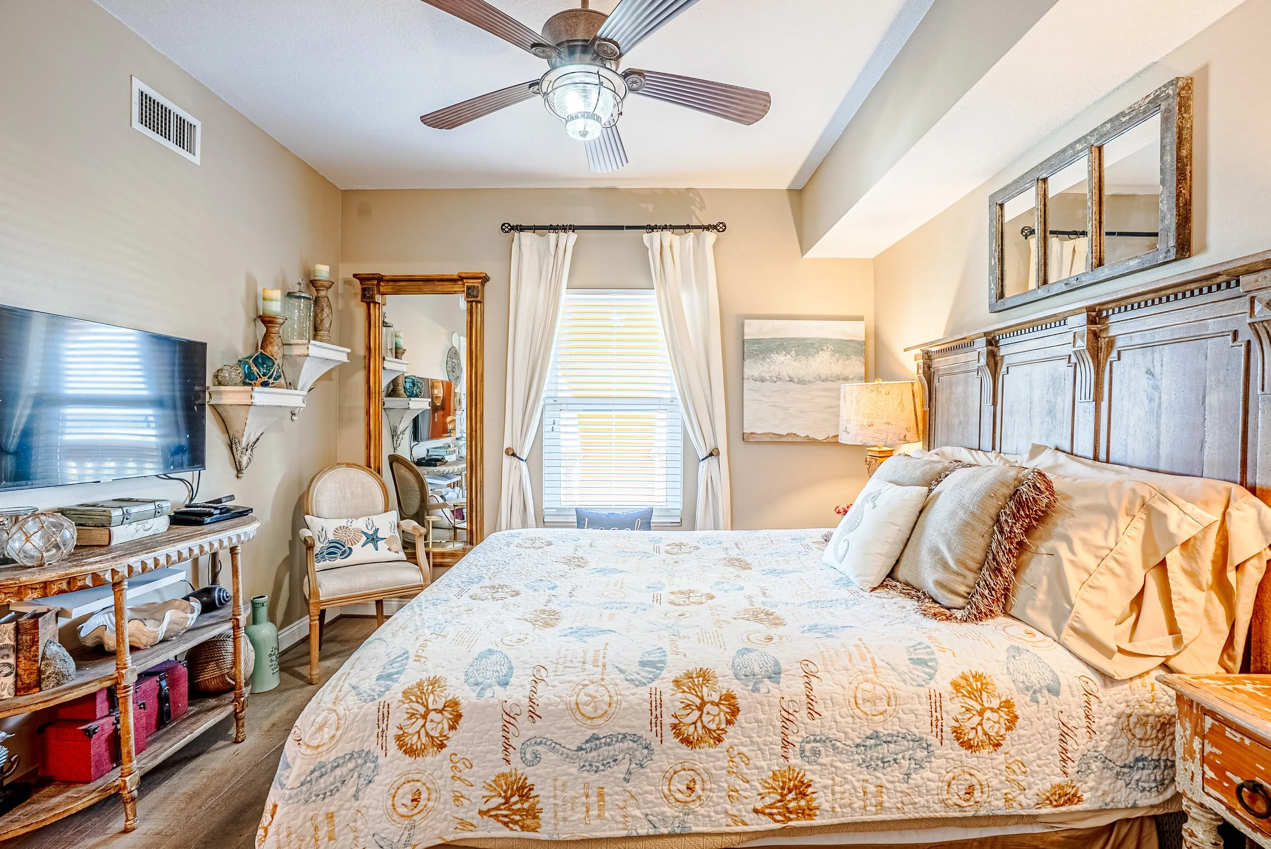 A bedroom with a ceiling fan, a window with white curtains, a large bed with pillows and a quilt, a mirror, a TV, and decorative shelves with candles and ornaments.