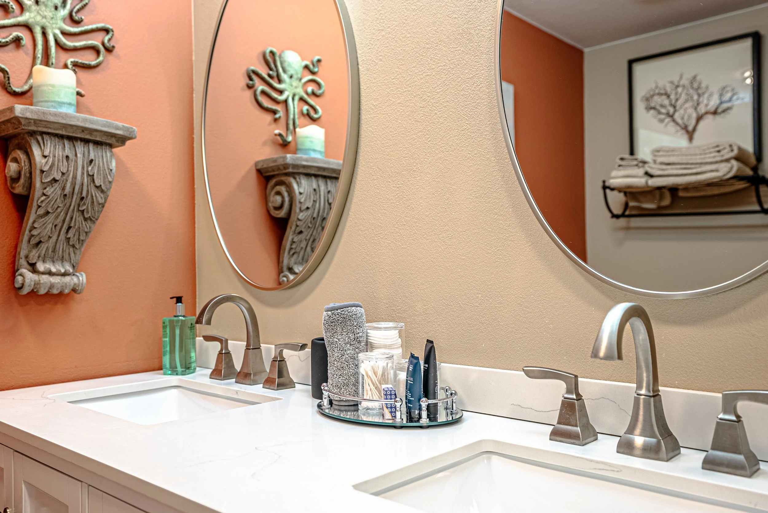 Bathroom sink with two mirrors, wooden wall decor, and shelves with folded towels and framed art in the background.