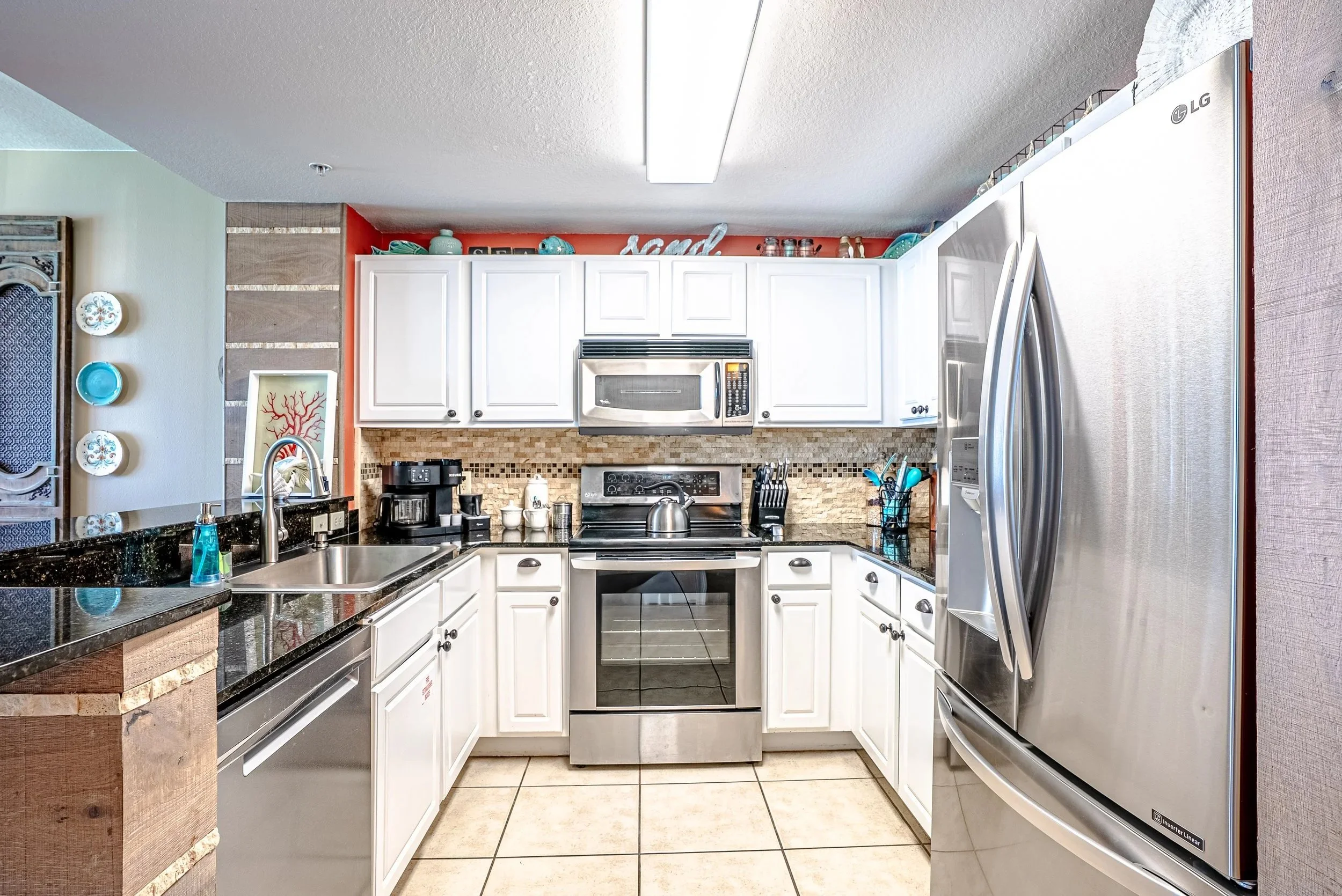 A kitchen with white cabinets, black granite countertops, a stainless steel refrigerator, and a white microwave oven above a stove. There are various kitchen utensils and appliances on the counters. The wall behind the stove has a beige tile backspla