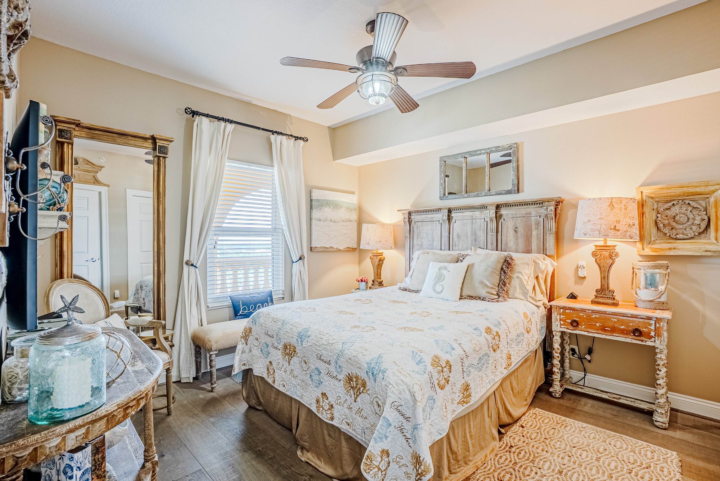 A cozy bedroom with a beige and white color scheme, featuring a large bed with beach-themed bedding, a wooden headboard, bedside lamps, a mirror above the headboard, and a window with white curtains letting in natural light.