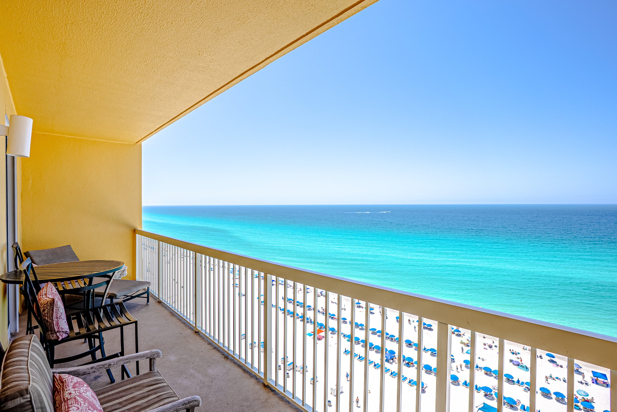 Balcony with outdoor chairs and pillows overlooking a beach with umbrellas and people, ocean and clear blue sky in the background.