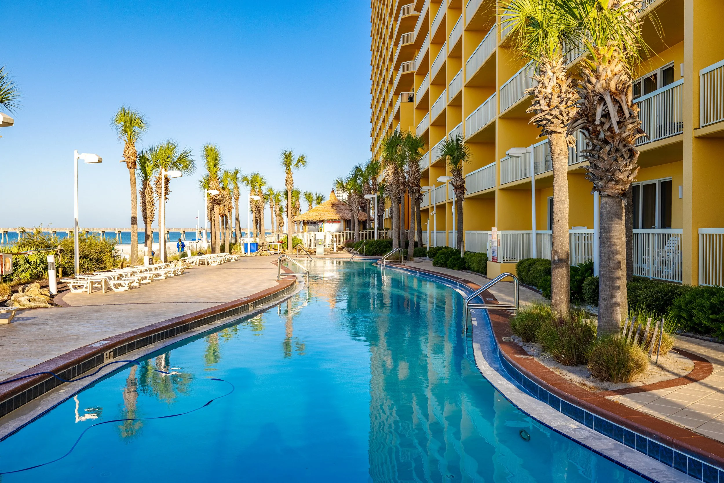 View of a hotel poolside area with palm trees, lounge chairs, and a yellow hotel building with balconies, under a clear blue sky.