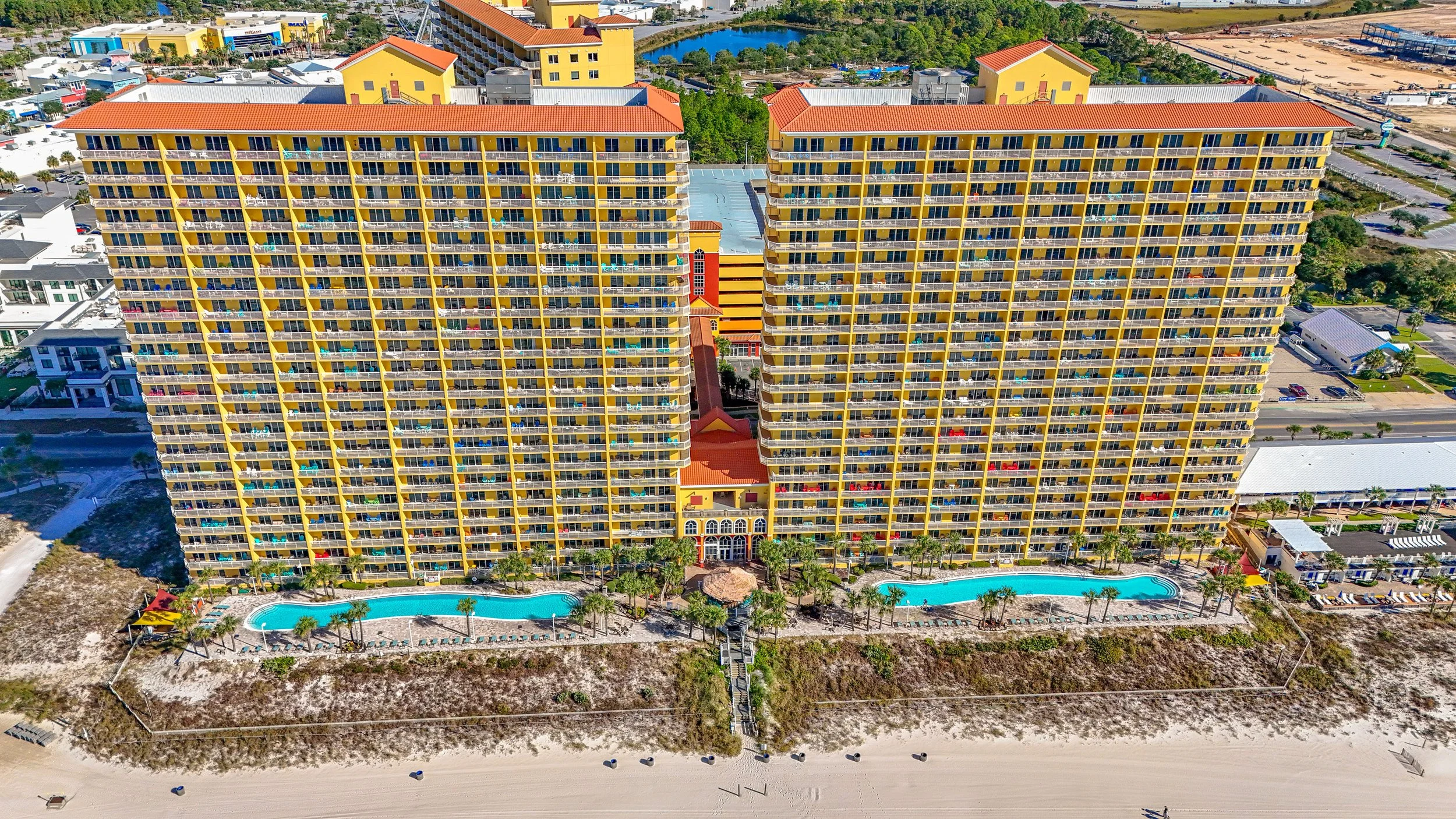An aerial view of two tall, yellow beachfront hotel buildings with balconies, a large swimming pool, and a sandy beach with lounge chairs and umbrellas at the bottom of the image. There are other buildings, roads, and green areas surrounding the hote