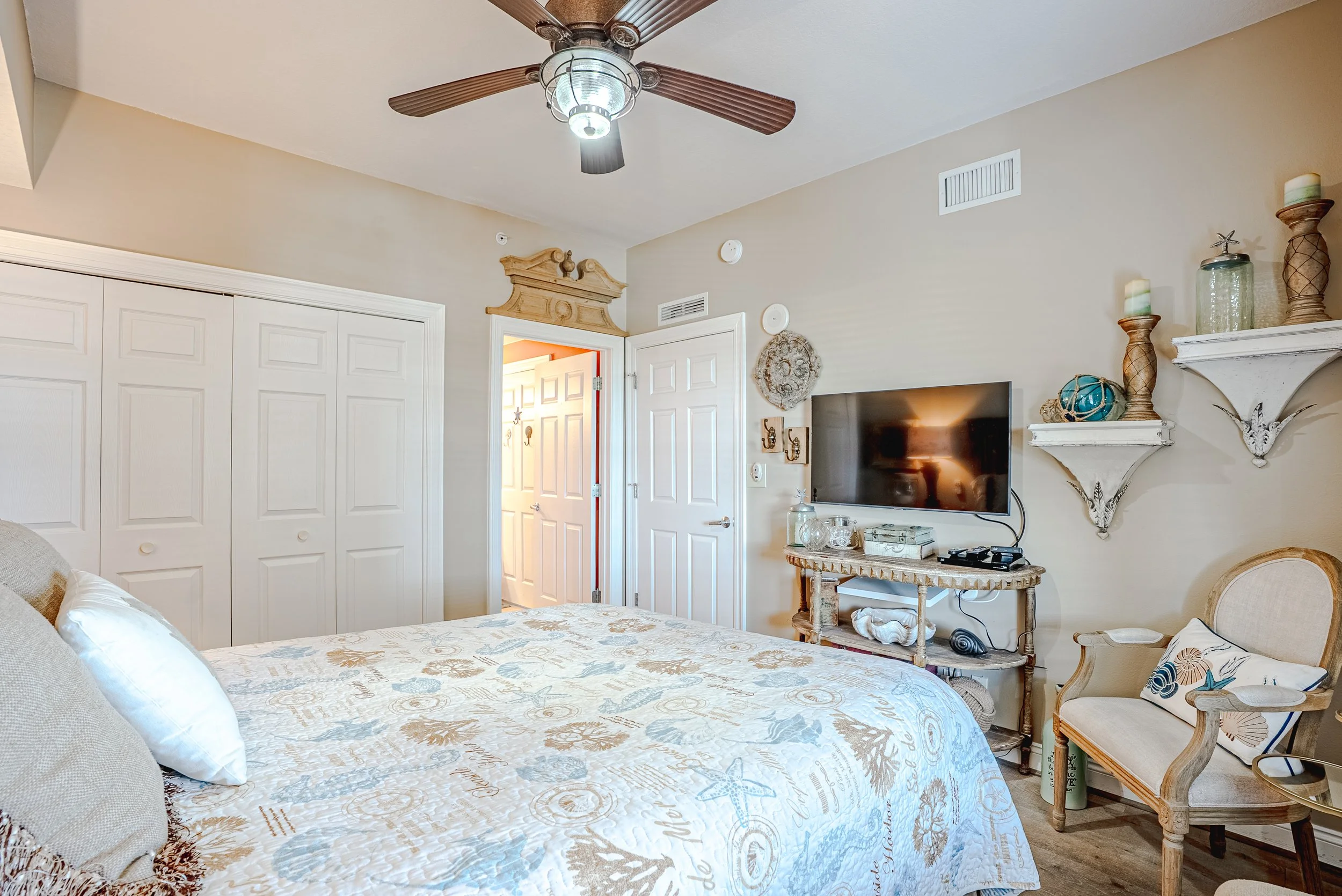 A bedroom with a large bed, a flat-screen TV, and various decorative items on shelves and tables. The room has light-colored walls, white closet doors, and a ceiling fan with wooden blades and a light fixture.