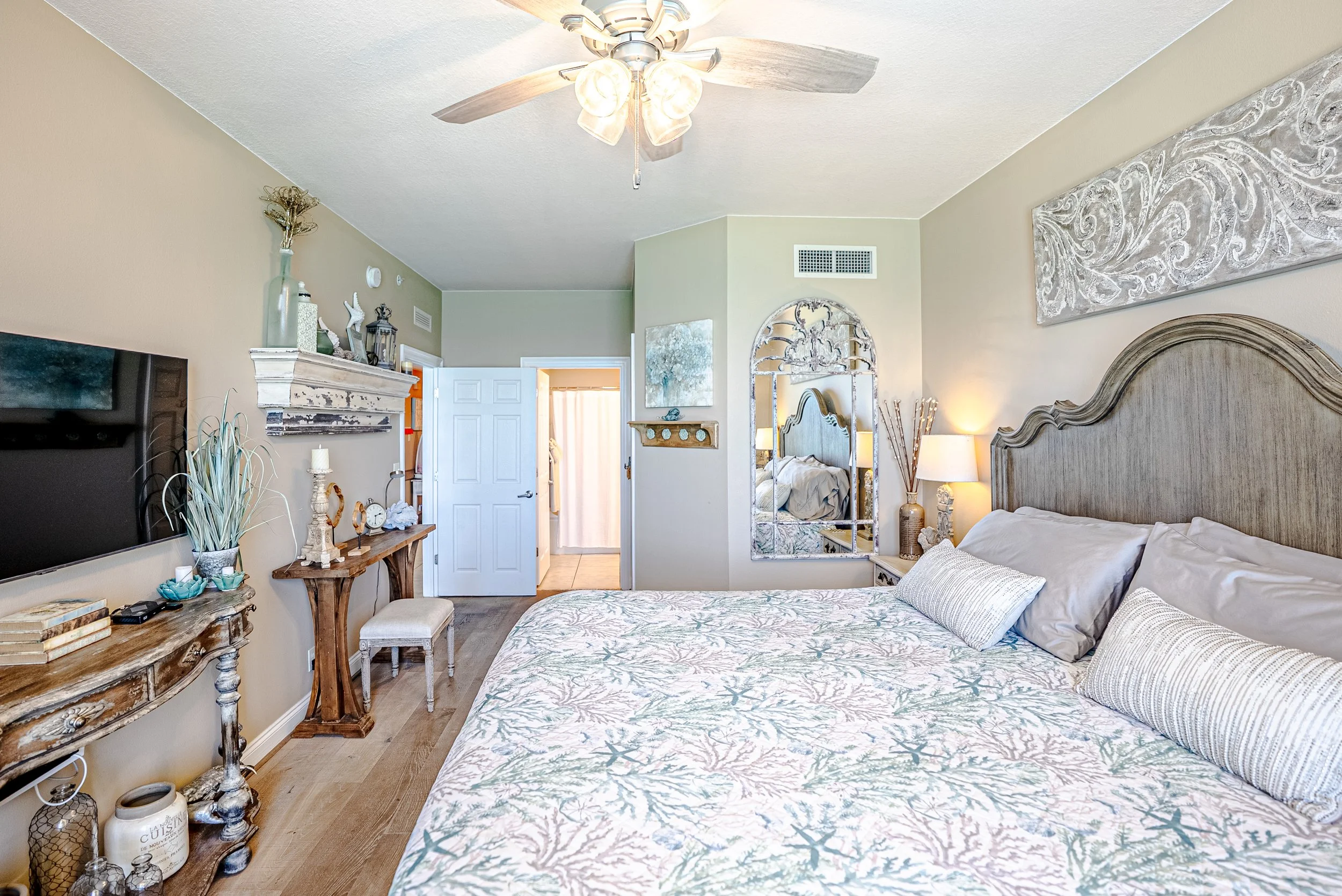 A cozy bedroom with a large bed covered in a nautical-themed bedspread and pillows. There is a rustic wooden dresser with decorative items, a wall-mounted mirror, and wall art. The room has a ceiling fan with lights, a small bench at the foot of the 