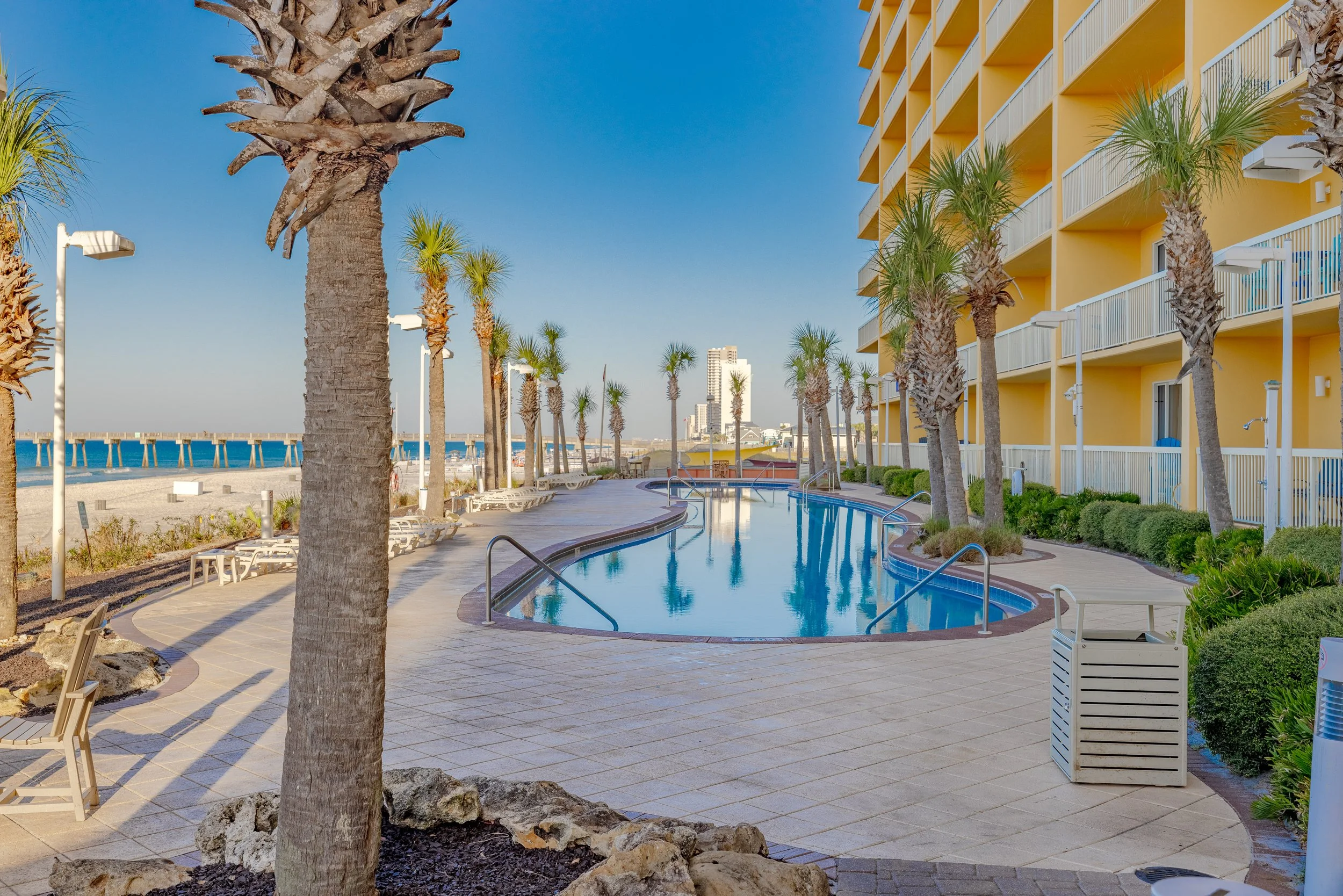 An outdoor scene of a hotel pool area with palm trees, lounge chairs, a pathway, a building with yellow walls and balconies, and a glimpse of the ocean and pier in the background under a clear blue sky.