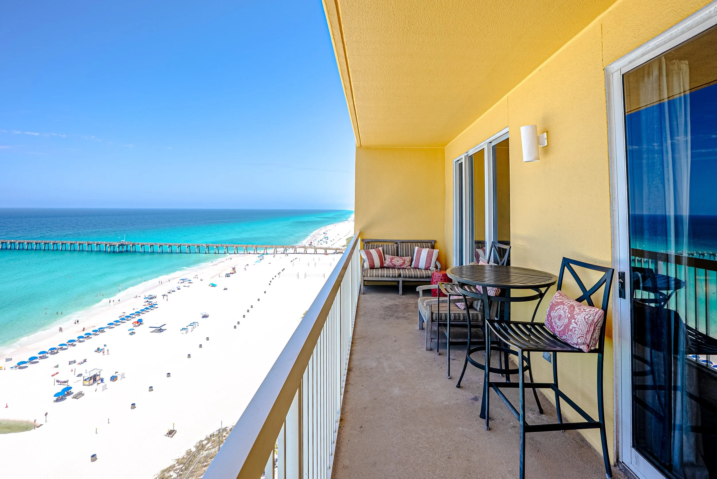 Balcony view of a beach with umbrellas and people, overlooking the ocean and a pier, with a yellow building wall and patio furniture in the foreground.