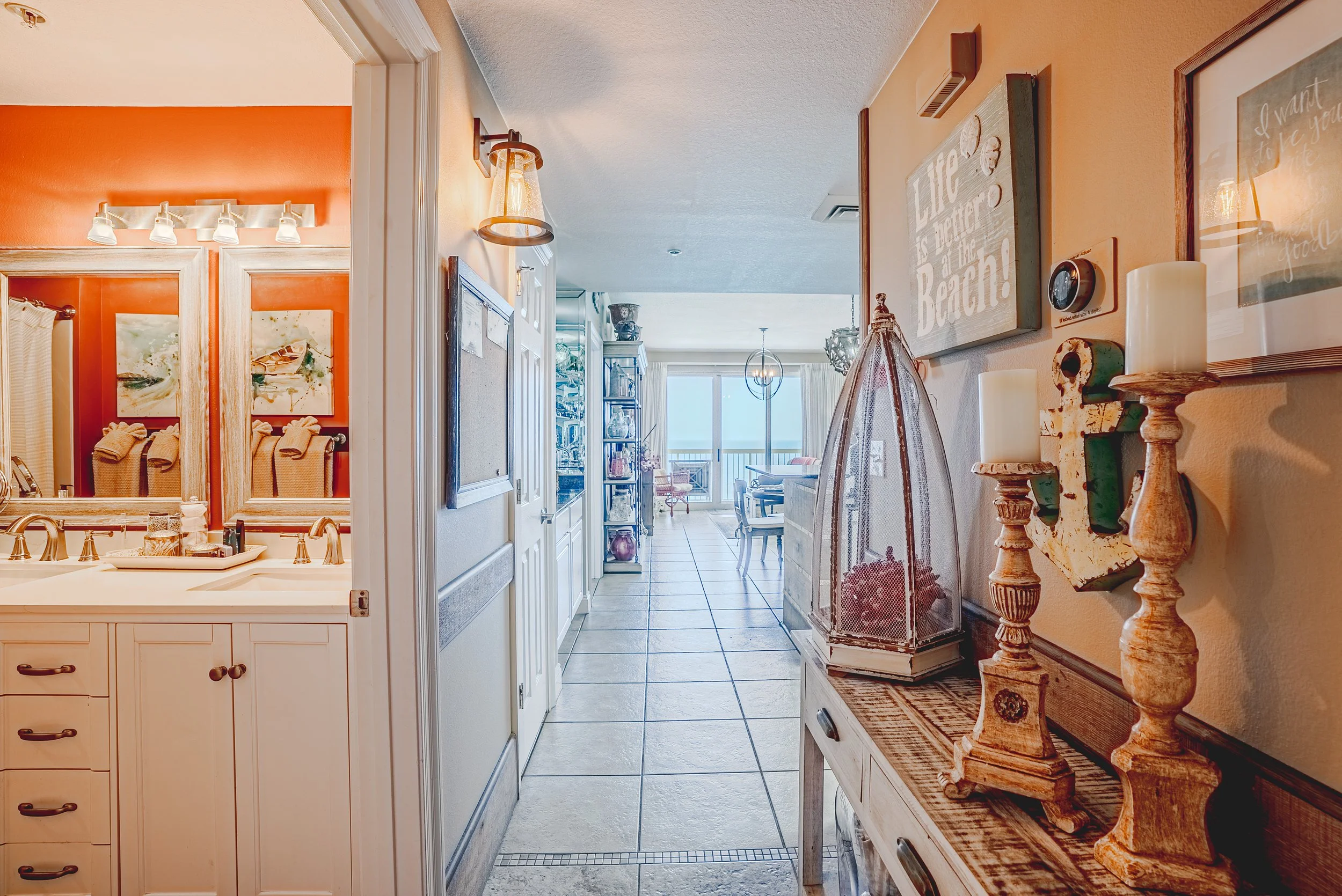 Interior of a home showing a hallway with a view into a dining area, decorated with lamps, paintings, and various home decor items, leading to a balcony.