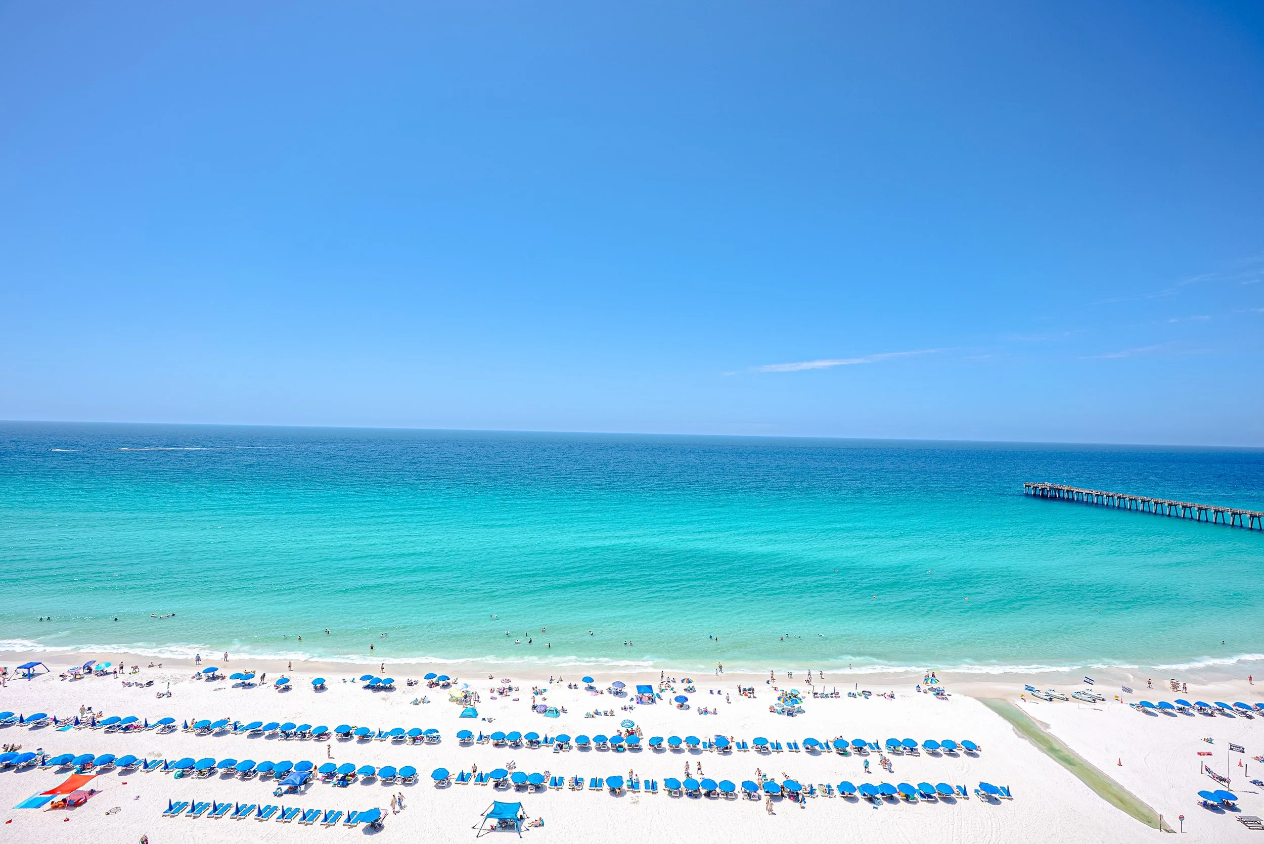 A sunny beach with blue umbrellas and people swimming in the ocean. A pier extends into the water on the right side.