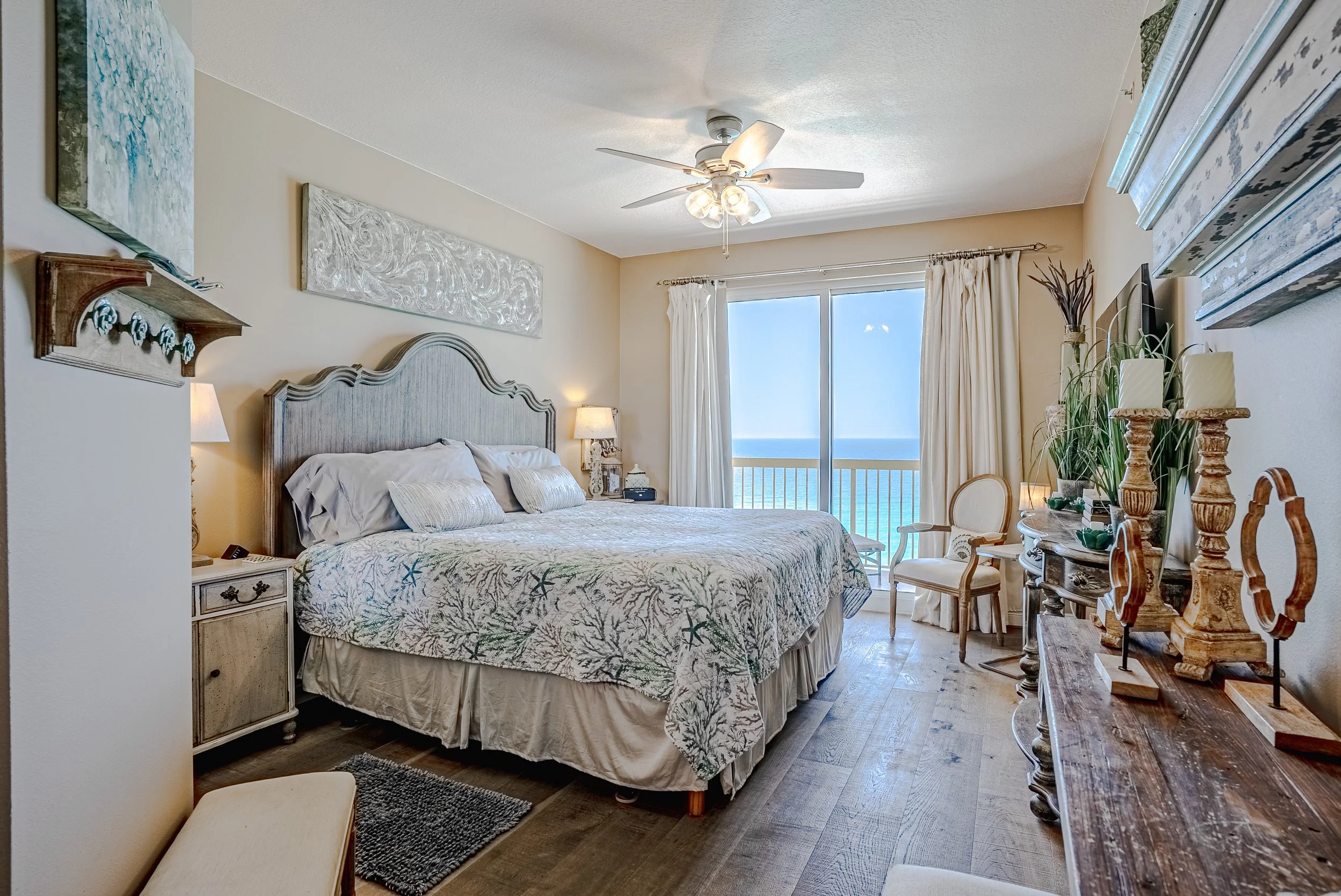A bedroom with a large bed, ocean view through sliding glass doors, beige walls, ceiling fan, and tropical decor including paintings, candles, and plants.