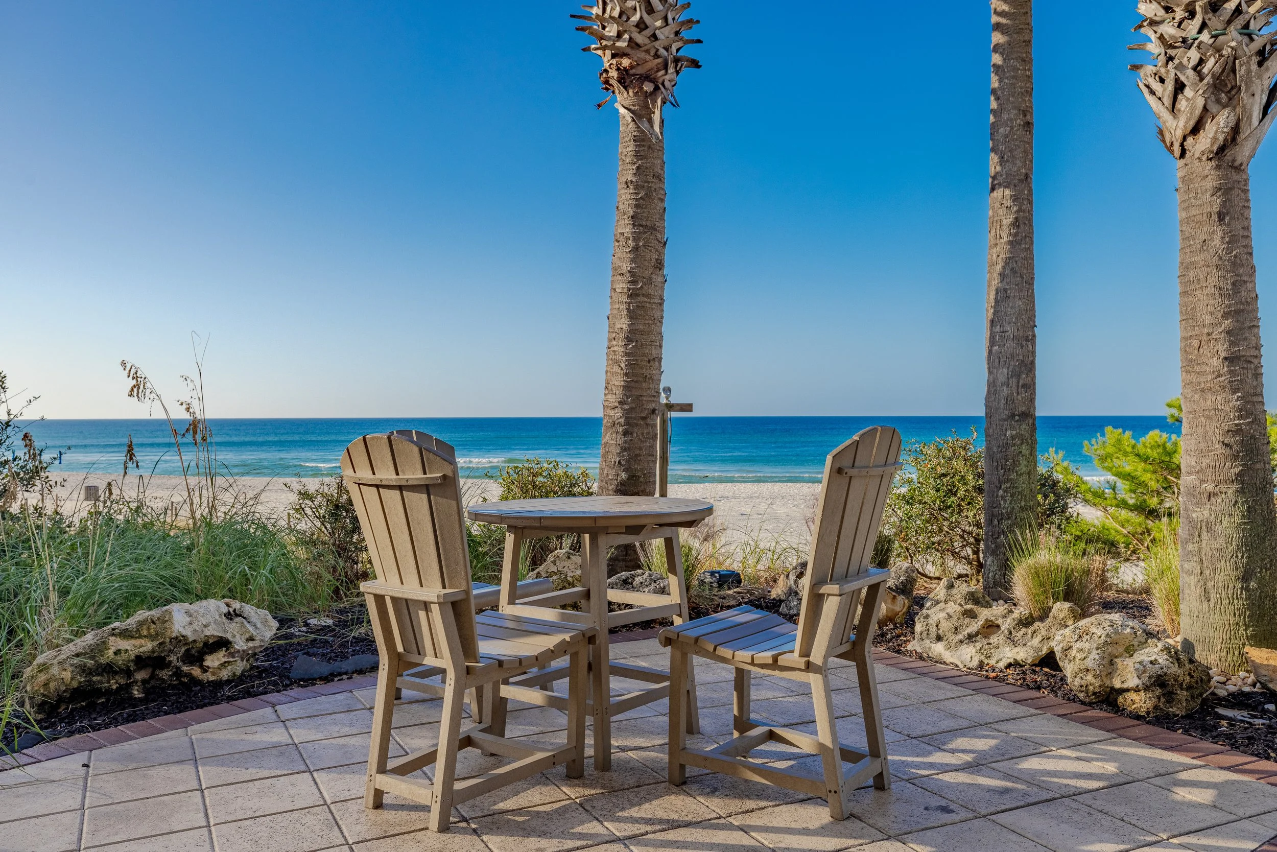 A beachside scene with four palm trees, a clear blue sky, and the ocean in the background. In the foreground, there are two wooden Adirondack chairs and a small round table on a tiled patio, surrounded by rocks and beach vegetation.