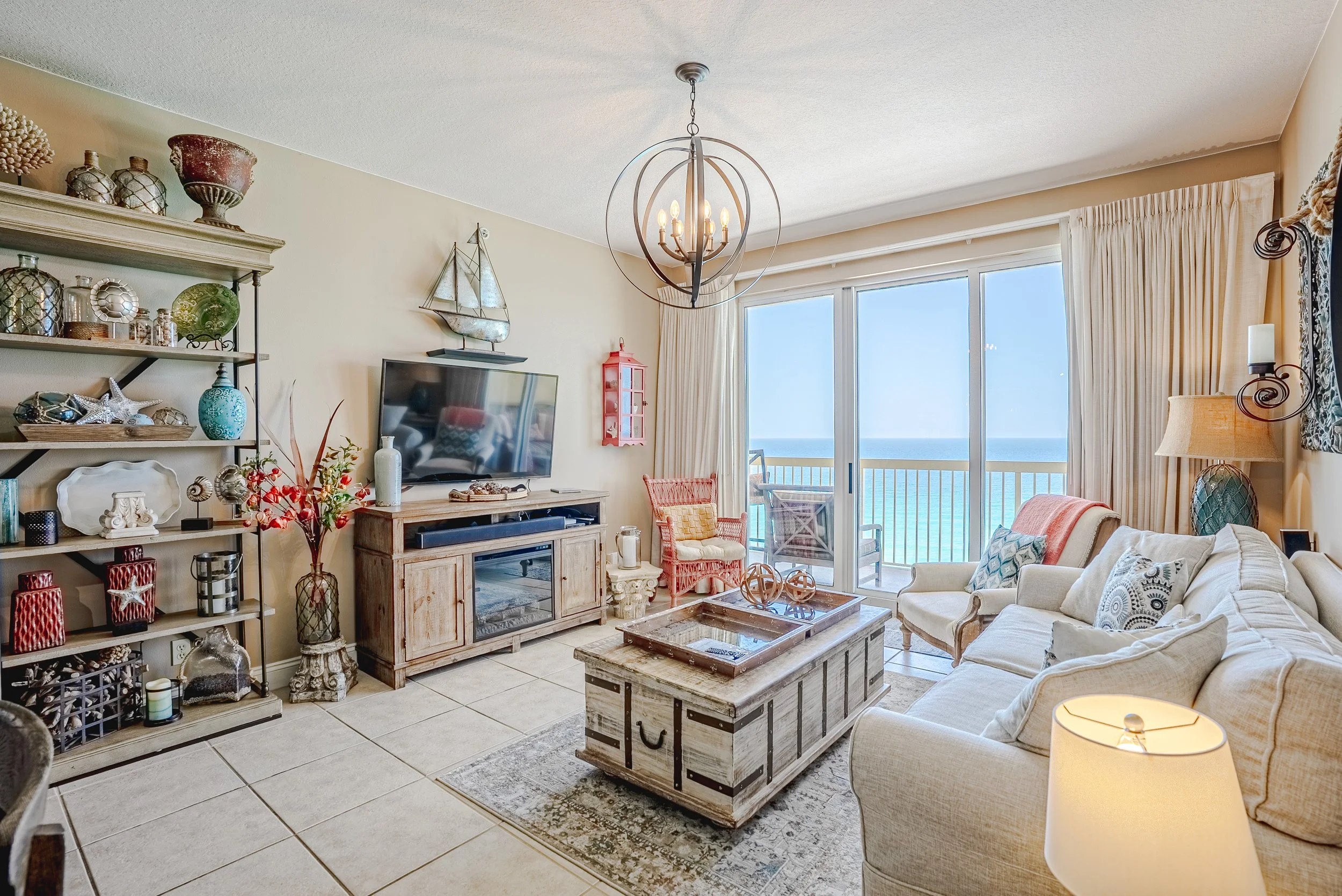 Living room with beige walls, white curtains, a beige sofa, a wooden coffee table, a TV on a wooden stand, a chandelier, a patterned rug, and large glass doors leading to a balcony with a view of the ocean.