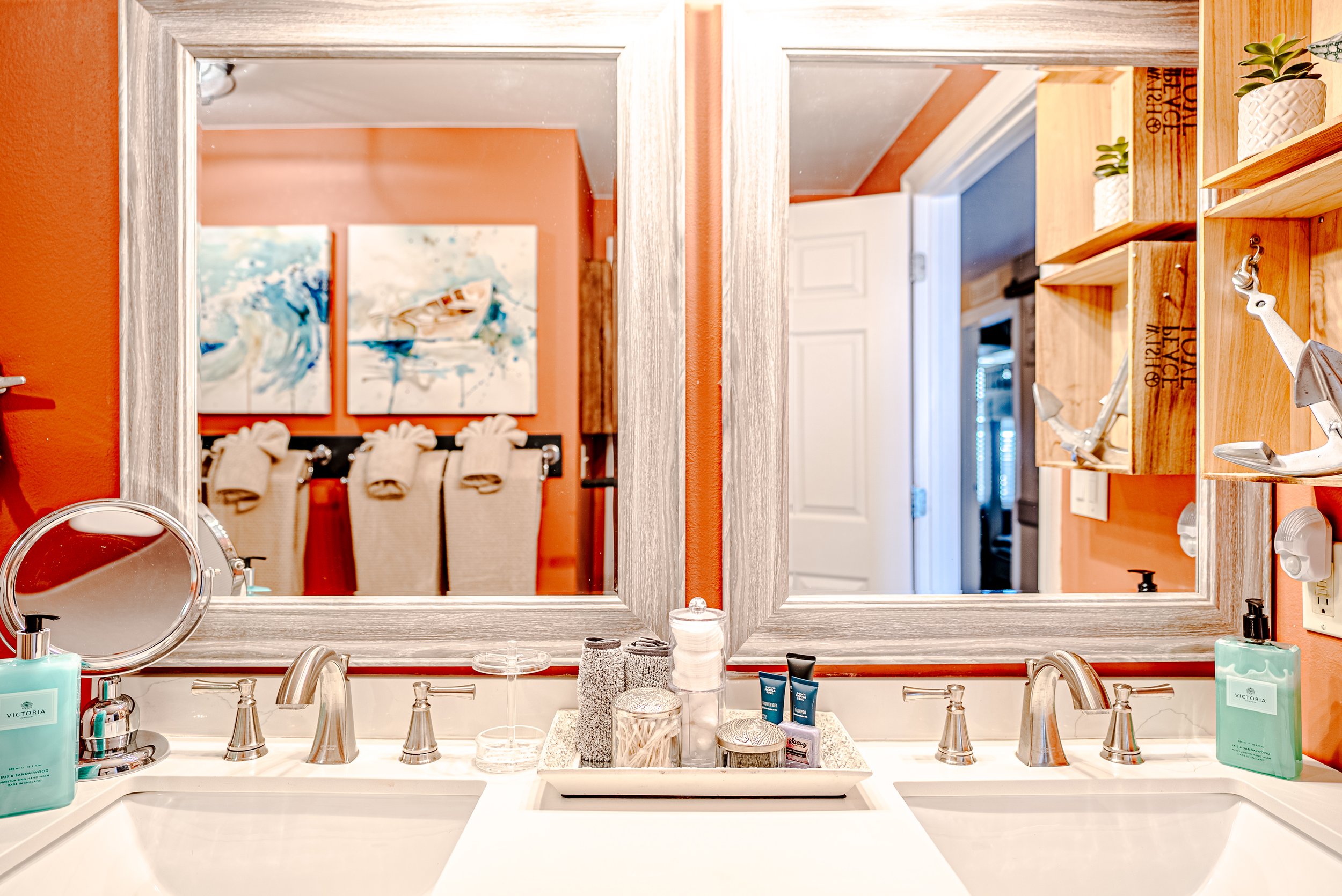 A bathroom vanity mirror reflecting a bathroom with orange walls, towels, artwork, and shelves with small potted plants. The sink has various toiletries and a silver faucet.