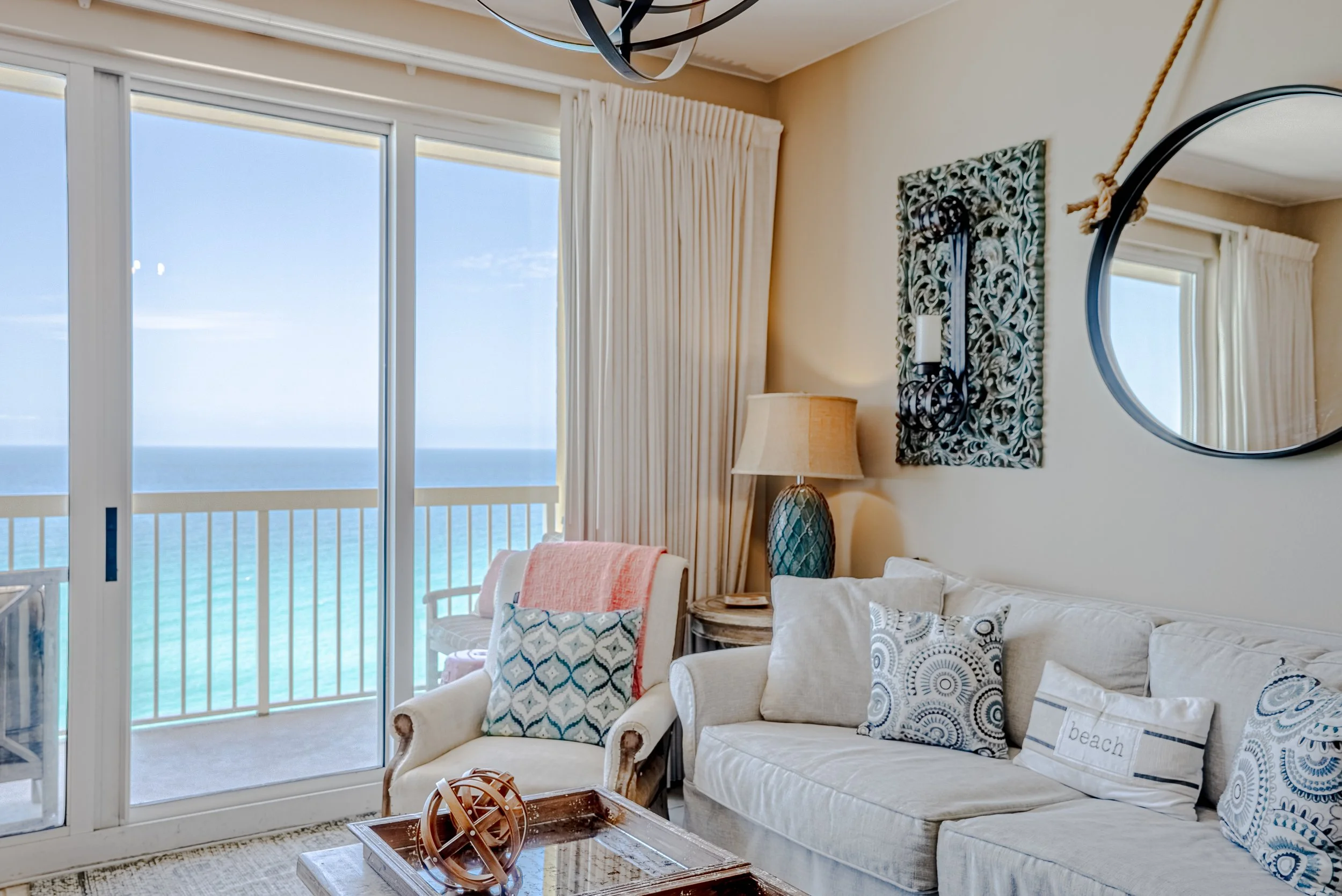 Living room with cream-colored sofa and armchair, decorative pillows, artwork on wall, round mirror, table lamp, sliding glass door with curtains, ocean view outside.