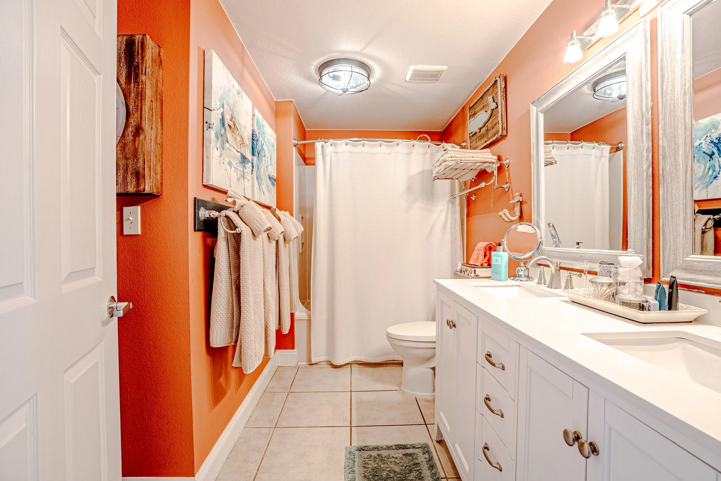 A bathroom with orange walls, a white vanity with a sink, mirror, and toiletries, beige tile floors, and a shower with a curtain. Towel racks and artwork decorate the space.