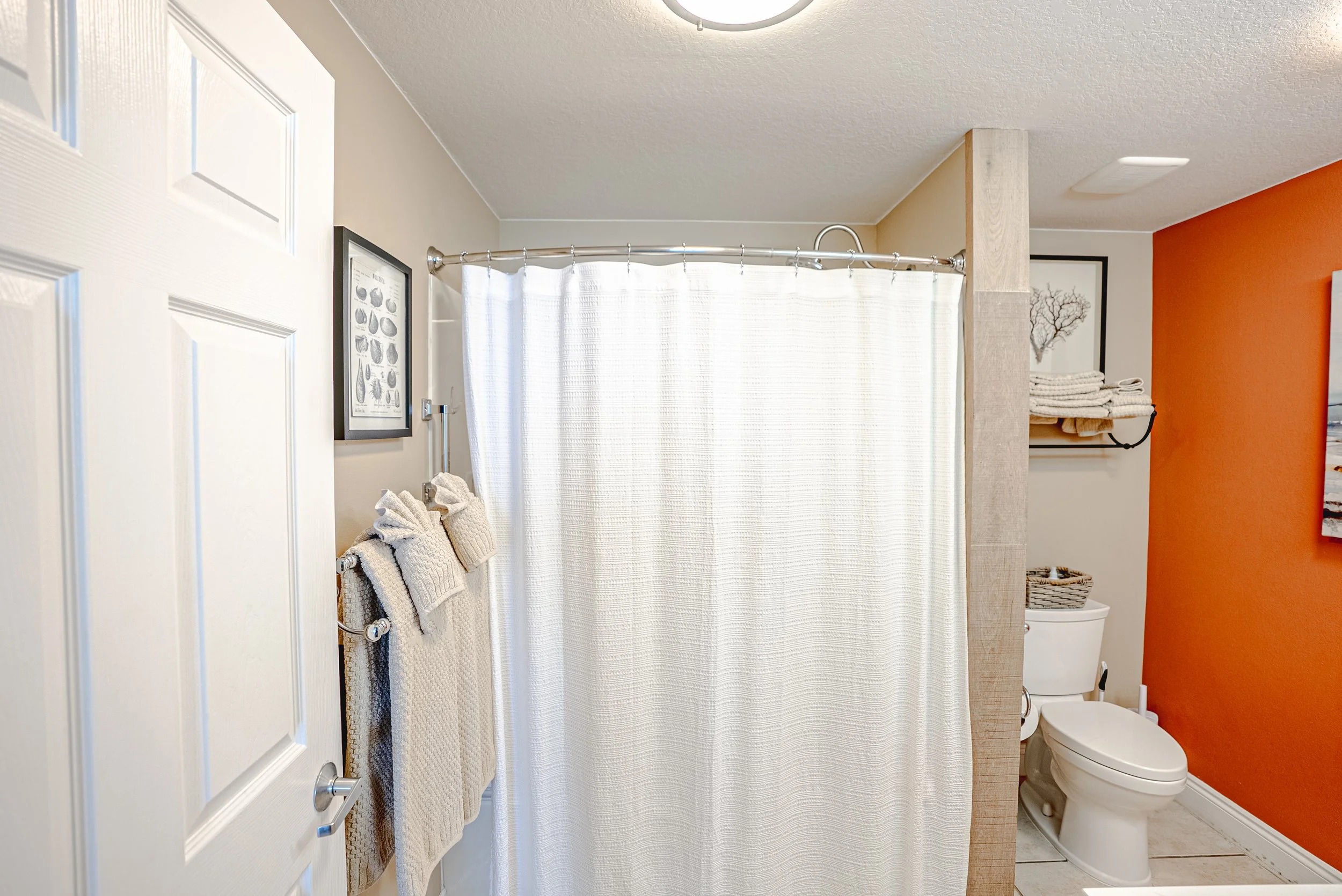 Bathroom with a white door, beige tiles, orange accent wall, a toilet, a shower with a curtain, and shelves with towels.
