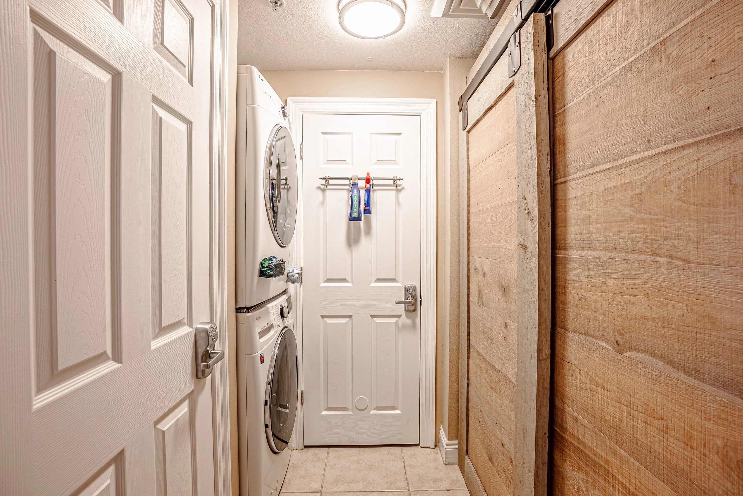 A laundry room with a stacked washer and dryer beside a white door, a towel rack with cleaning supplies hanging, and a wooden accent wall on the right.