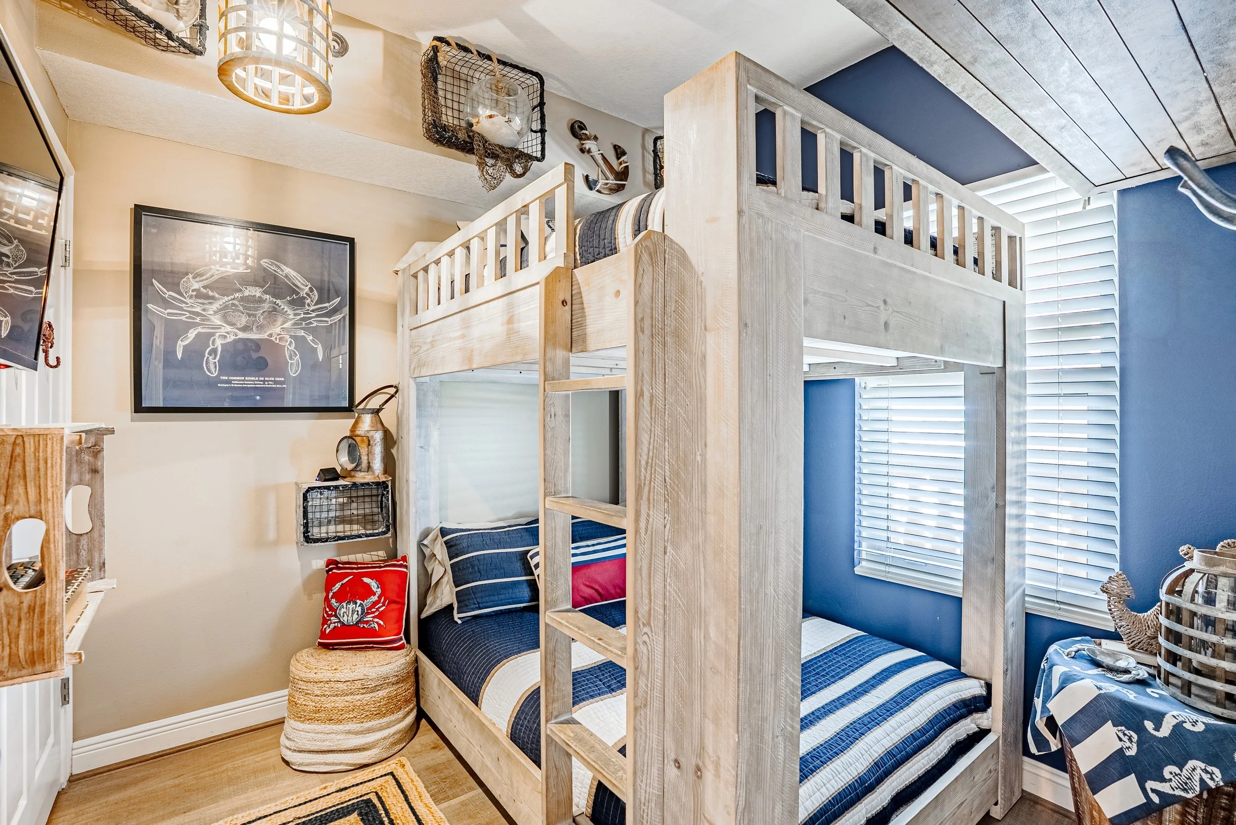 Bedroom with light wood bunk bed, striped bedding, blue and striped pillows, large windows with white blinds, nautical decor including an anchor and crab print, wicker ottoman, and various decorative items.