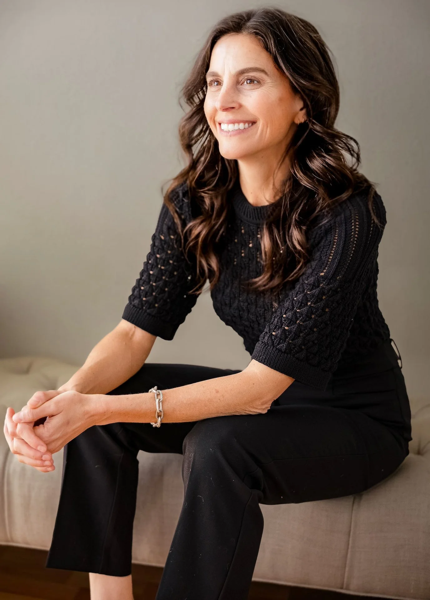 A woman with wavy brown hair, smiling, wearing a black knitted top and black pants, sitting on a beige couch indoors.