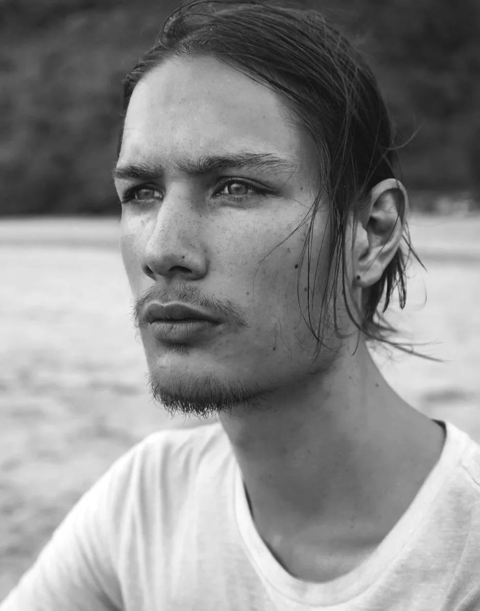 Close-up black and white portrait of a young man with long hair, some facial hair, and freckles, looking pensively into the distance, outdoors.