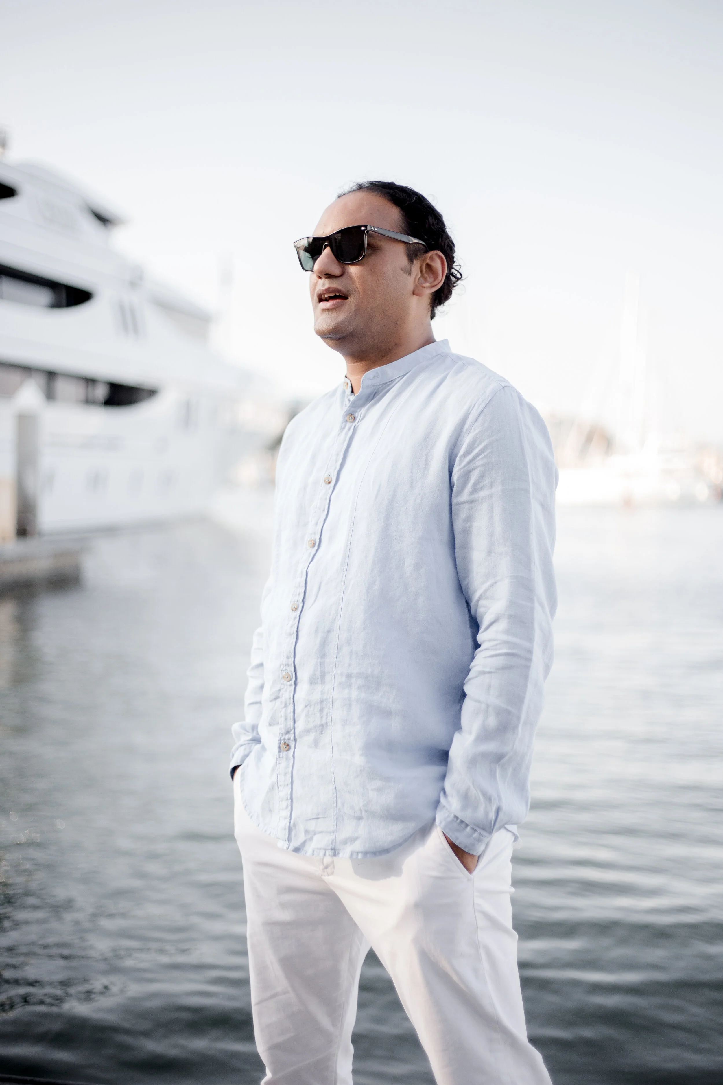 A man wearing dark sunglasses, a light blue button-up shirt, and white pants stands near a marina with yachts in the background, under a clear sky.