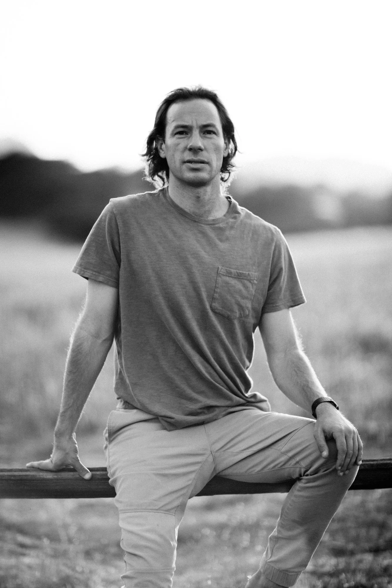 A black and white photo of a man with long hair sitting outdoors on a park bench, with trees and grass in the background.