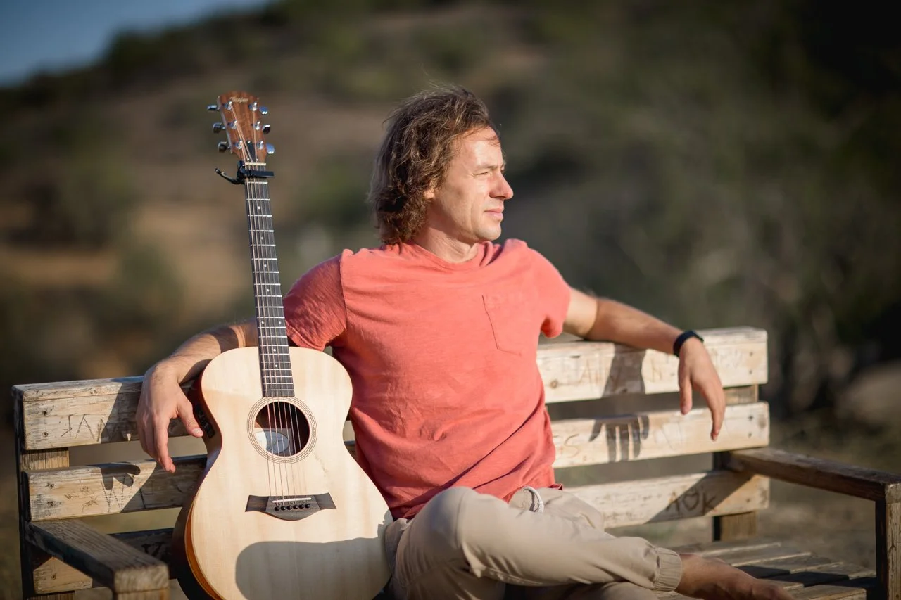 A man in a red t-shirt sitting on a wooden bench with a guitar resting beside him, outdoors during daytime with a blurred natural background.