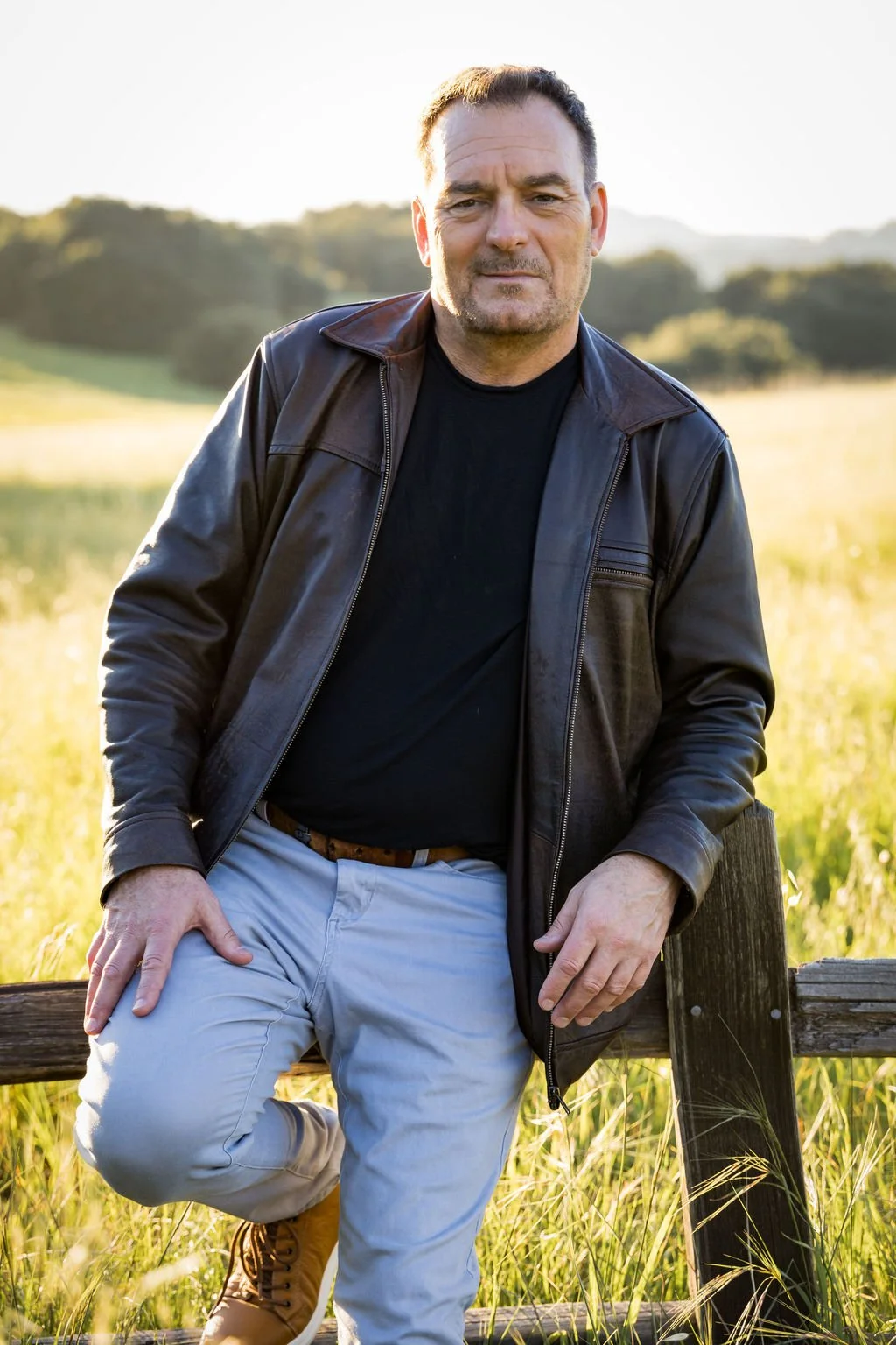 A man with short hair and stubble wearing a black shirt and black leather jacket, posing outdoors in a field with hills and water in the background during late afternoon sunlight.