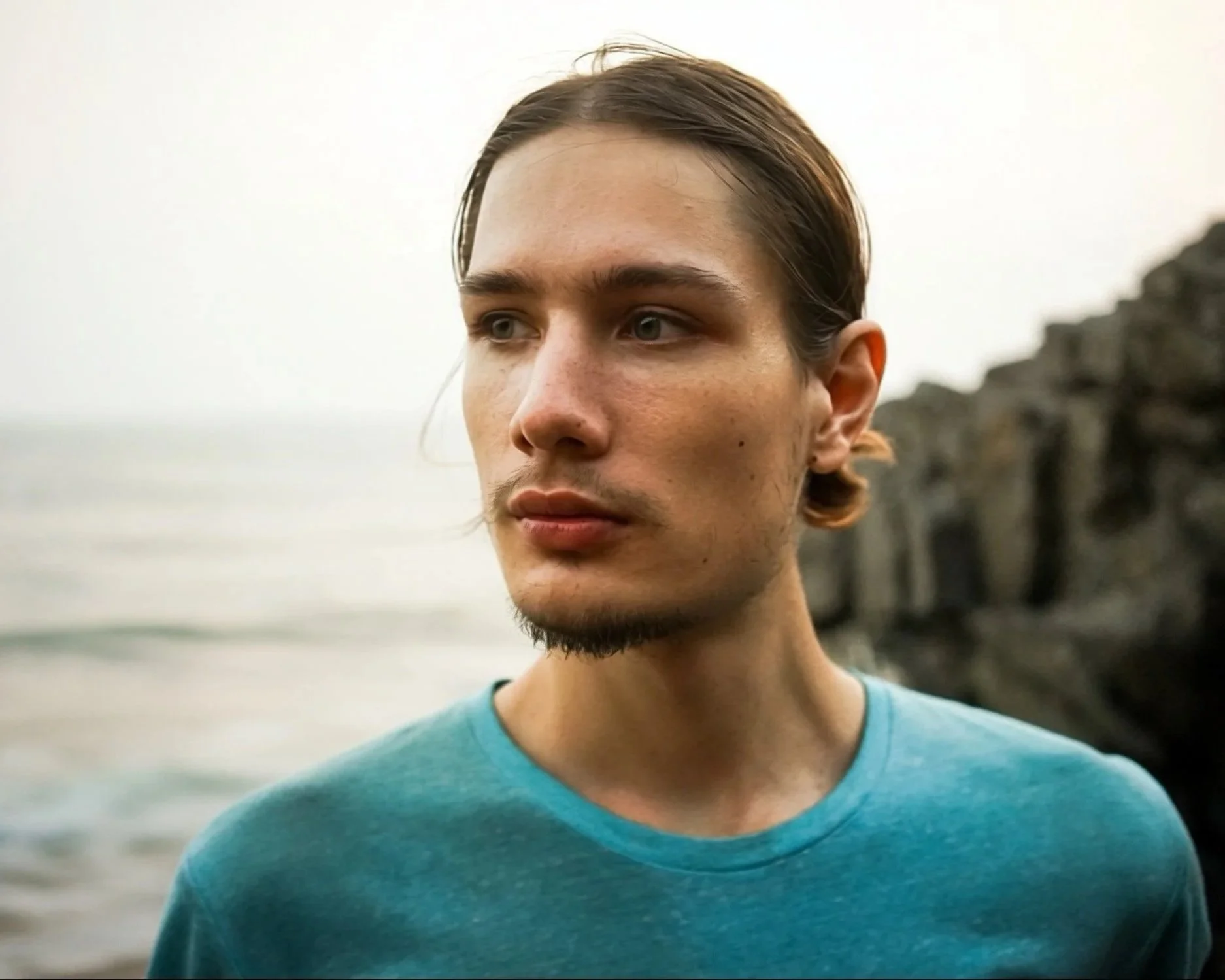 A young man with light brown hair, blue eyes, and a goatee wearing a teal t-shirt, standing outdoors near the coast with rocky cliffs and ocean in the background.