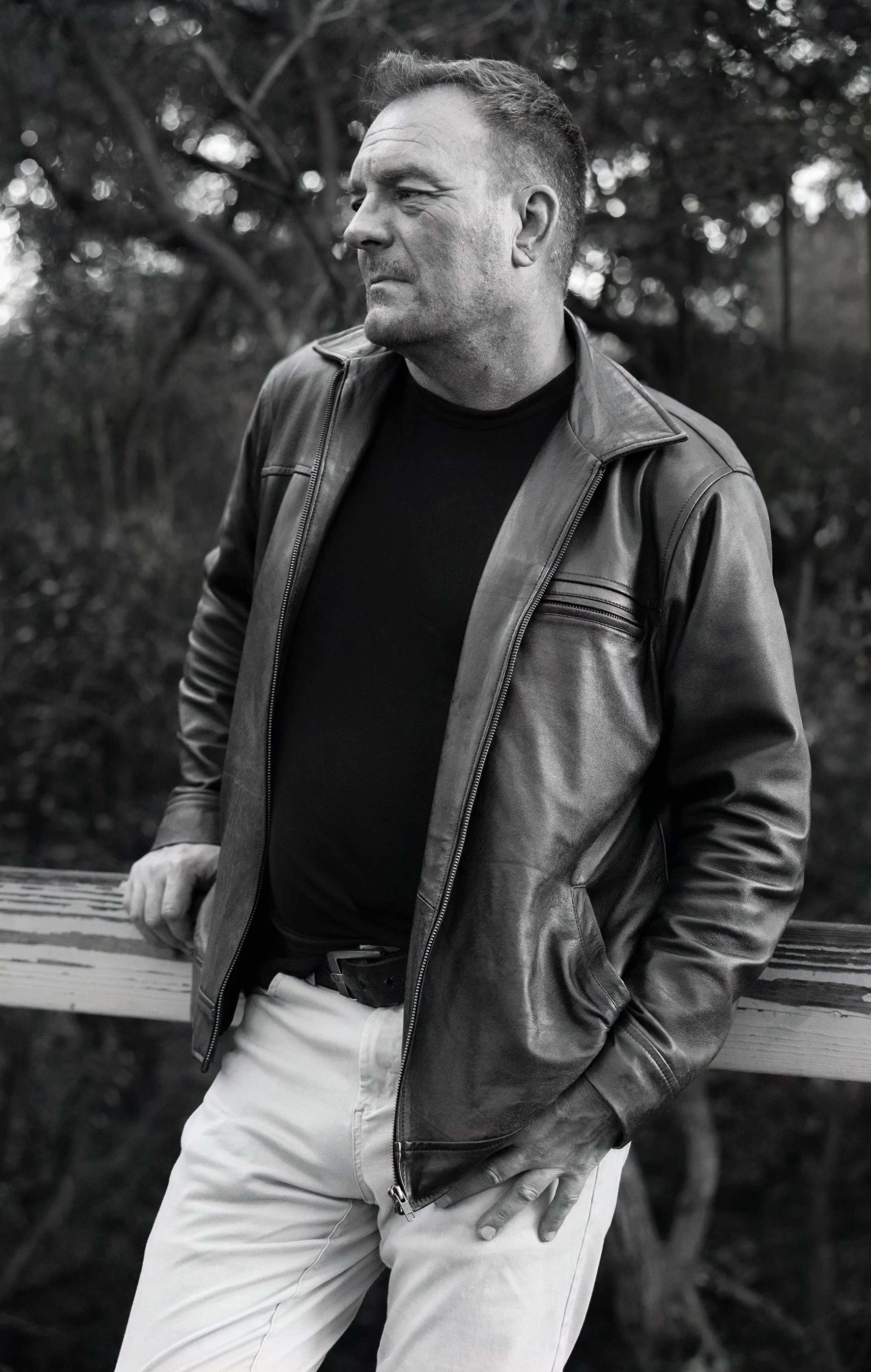 A black and white photo of a man wearing a leather jacket, black shirt, and light-colored pants, standing outdoors near a wooden railing with trees in the background.