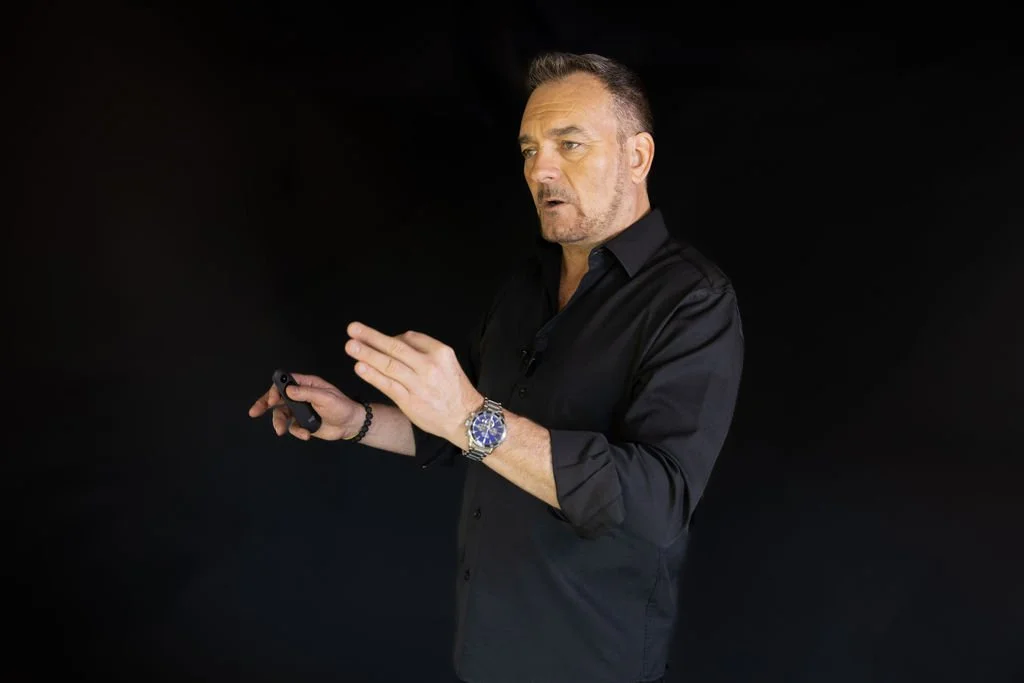 A man in a black shirt holding a remote control or laser pointer, standing against a black background.