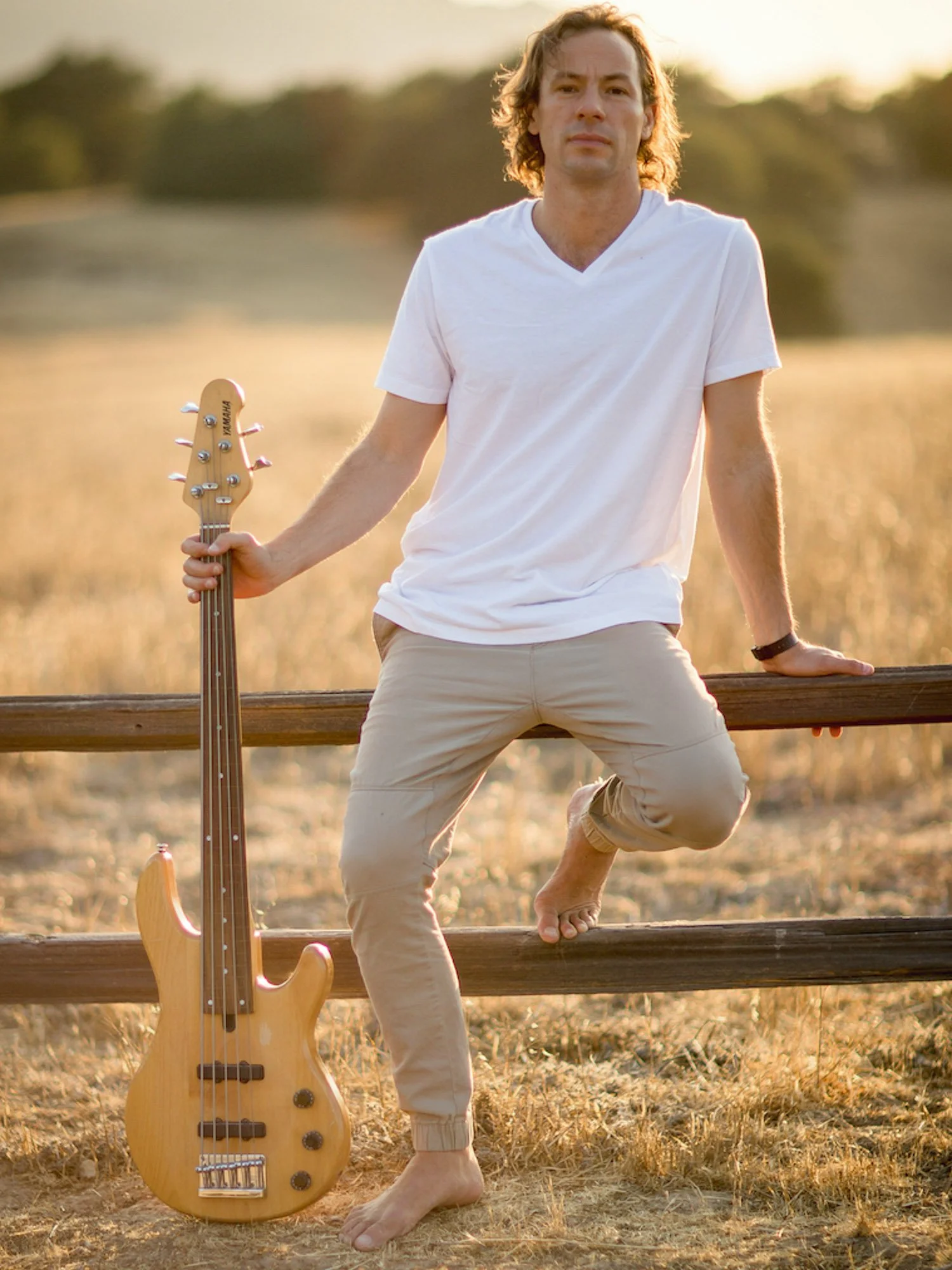 A man with wavy brown hair standing outdoors in a field at sunset, holding a bass guitar and leaning on a wooden railing, with one foot resting on the lower rail and the other foot on the ground.