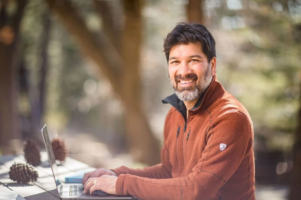 A man with dark hair and a beard smiling while sitting at an outdoor table with a laptop, wearing a brown jacket, with trees in the background.
