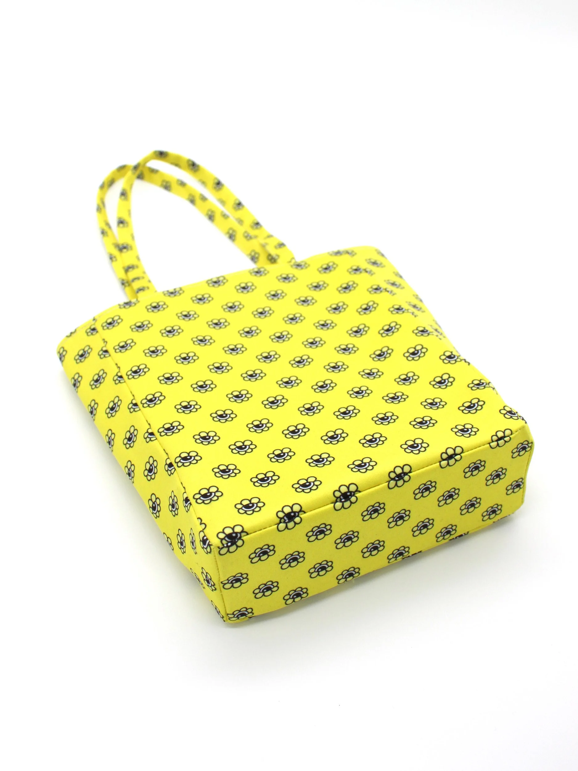 An angled view looking at the bottom and front of the Bri Foster Dolls yellow mini tote bag. The shot highlights the rectangular base and sturdy seam construction of the 'doll eyes' printed fabric.