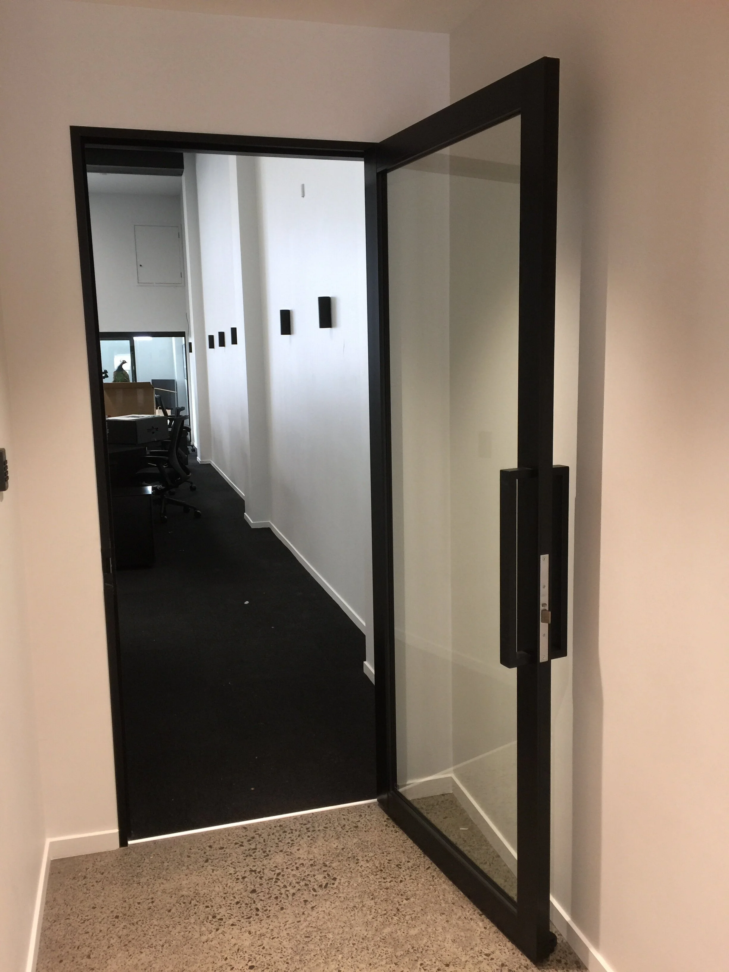 An open glass elevator door reveals a view into a modern office corridor with black carpet and white walls.