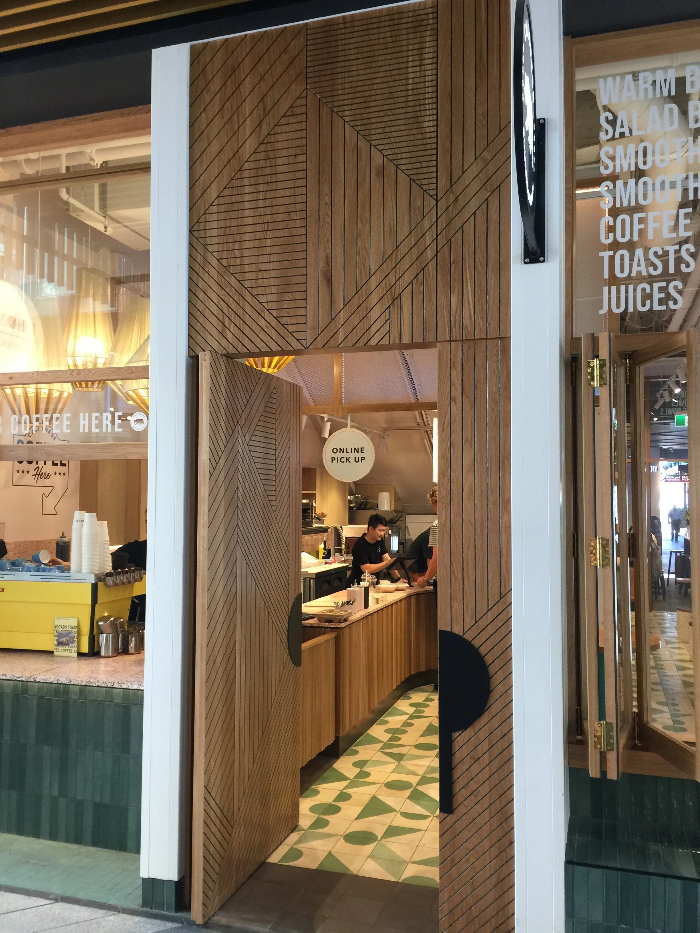 Interior of a modern cafe with wooden panel decor and a view through the open doorway showing staff working behind the counter. Signage indicates online pick up and menu offerings such as warm bread, salad, smoothies, coffee, toasts, and juices.