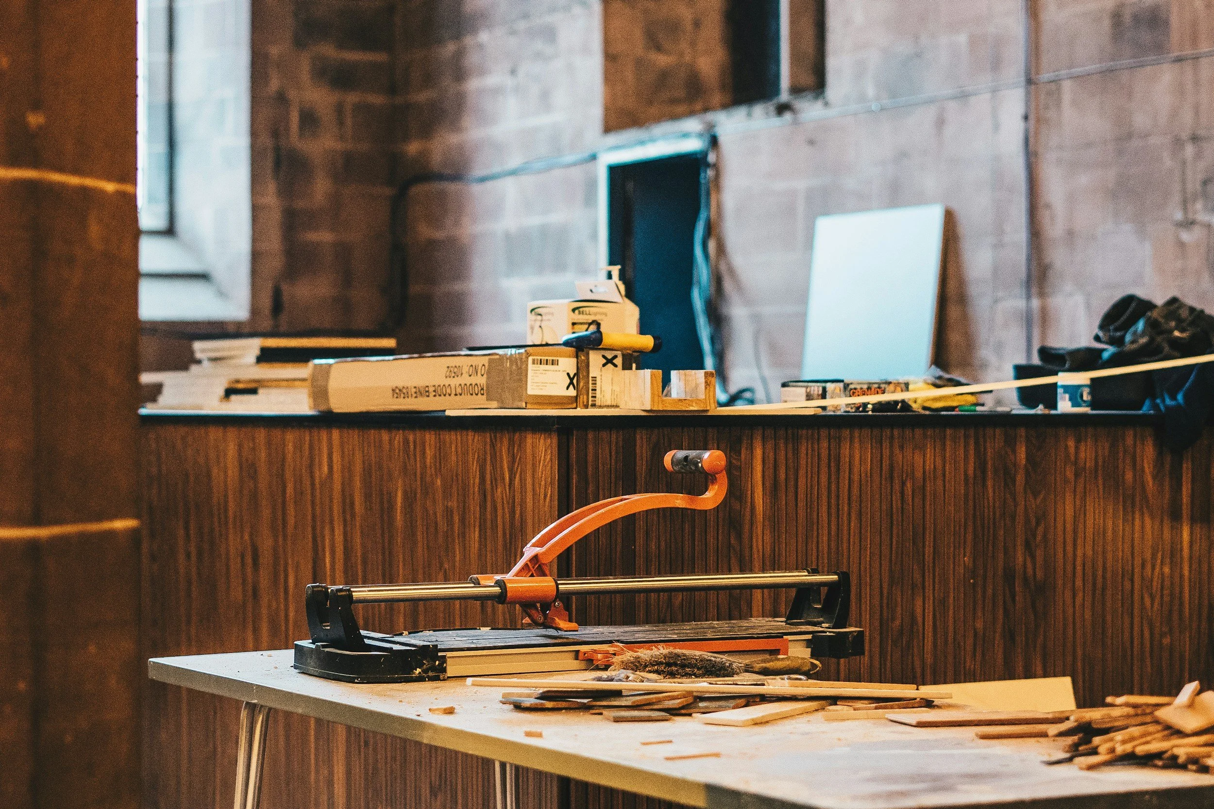 A cluttered workbench with a scroll saw, woodworking tools, and materials in a wood-paneled workshop.